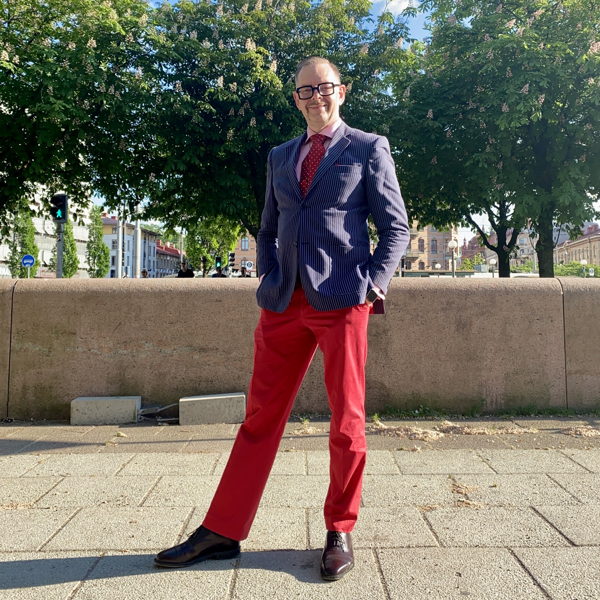 Man in a striped blazer and red trousers standing confidently outdoors with trees in the background.