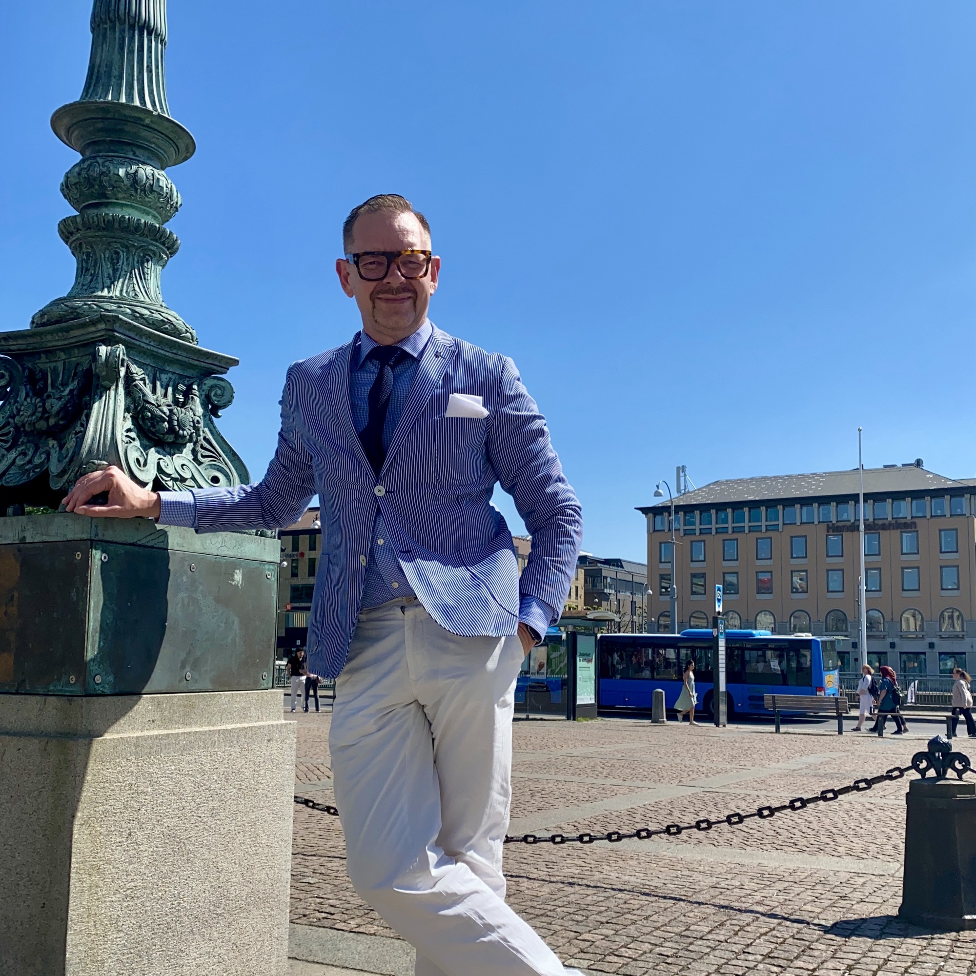Man in a light blue blazer and white trousers standing beside a decorative lamp post in an urban setting.