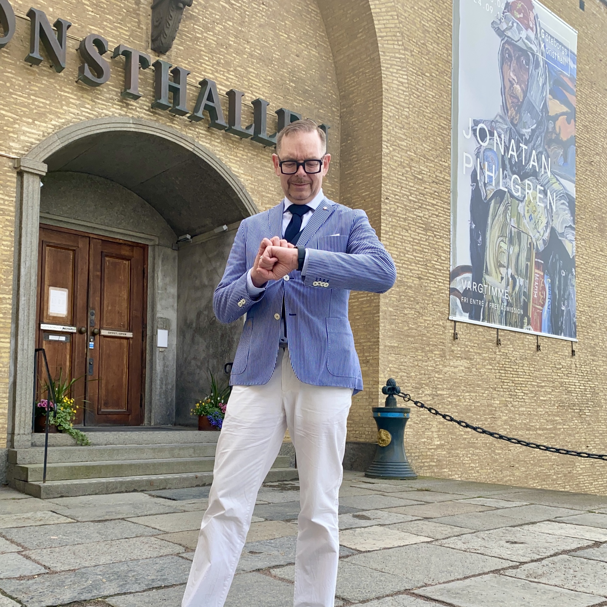 Man in a blue blazer and white trousers checking his watch outside a building with a large poster.