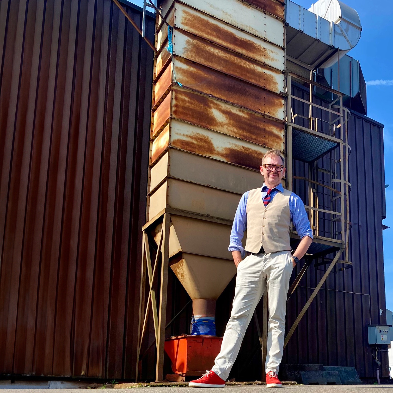 Man in a vest and tie standing confidently next to a large industrial structure with rusted metal surfaces.
