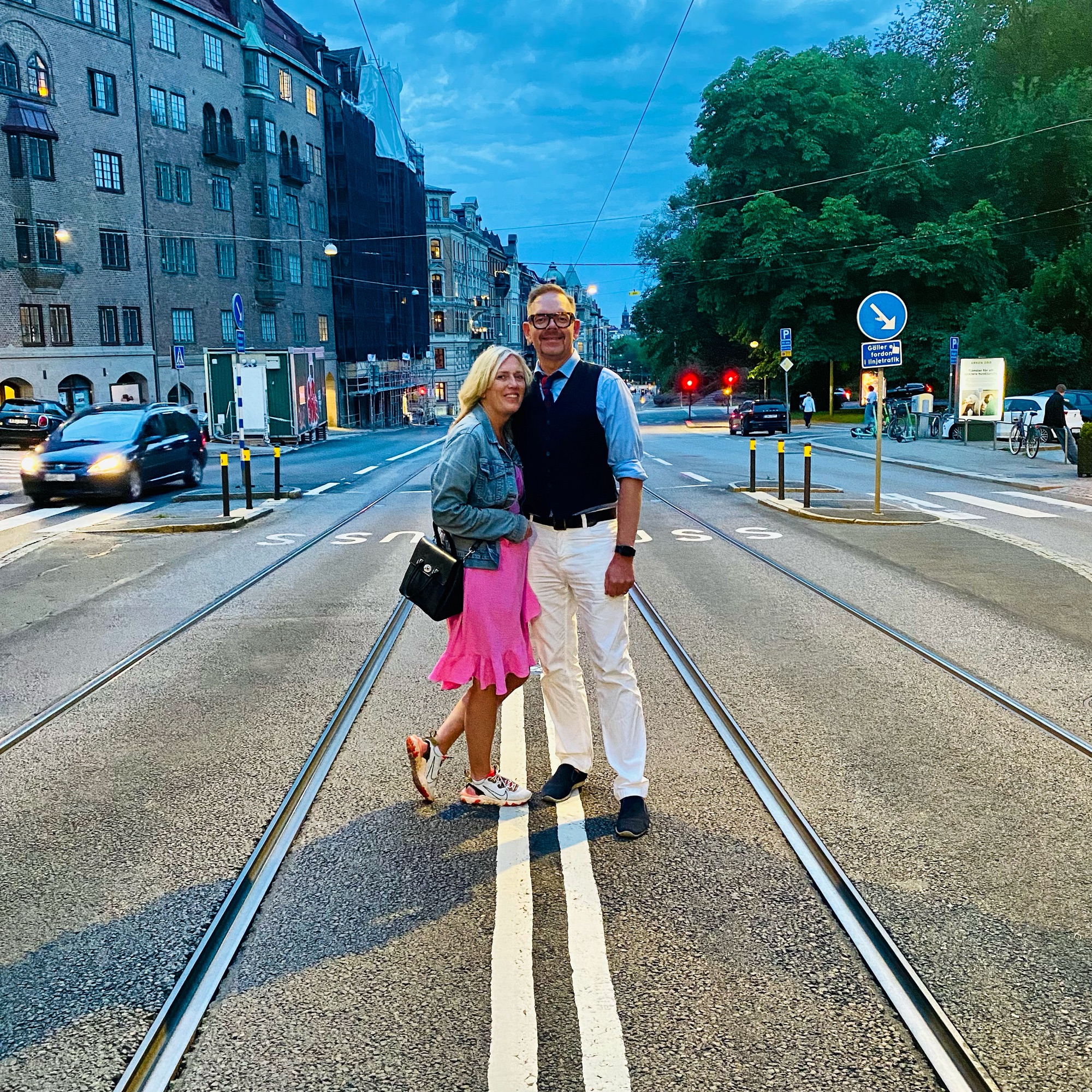 A couple standing on tram tracks in a city street during twilight, with buildings and trees in the background.
