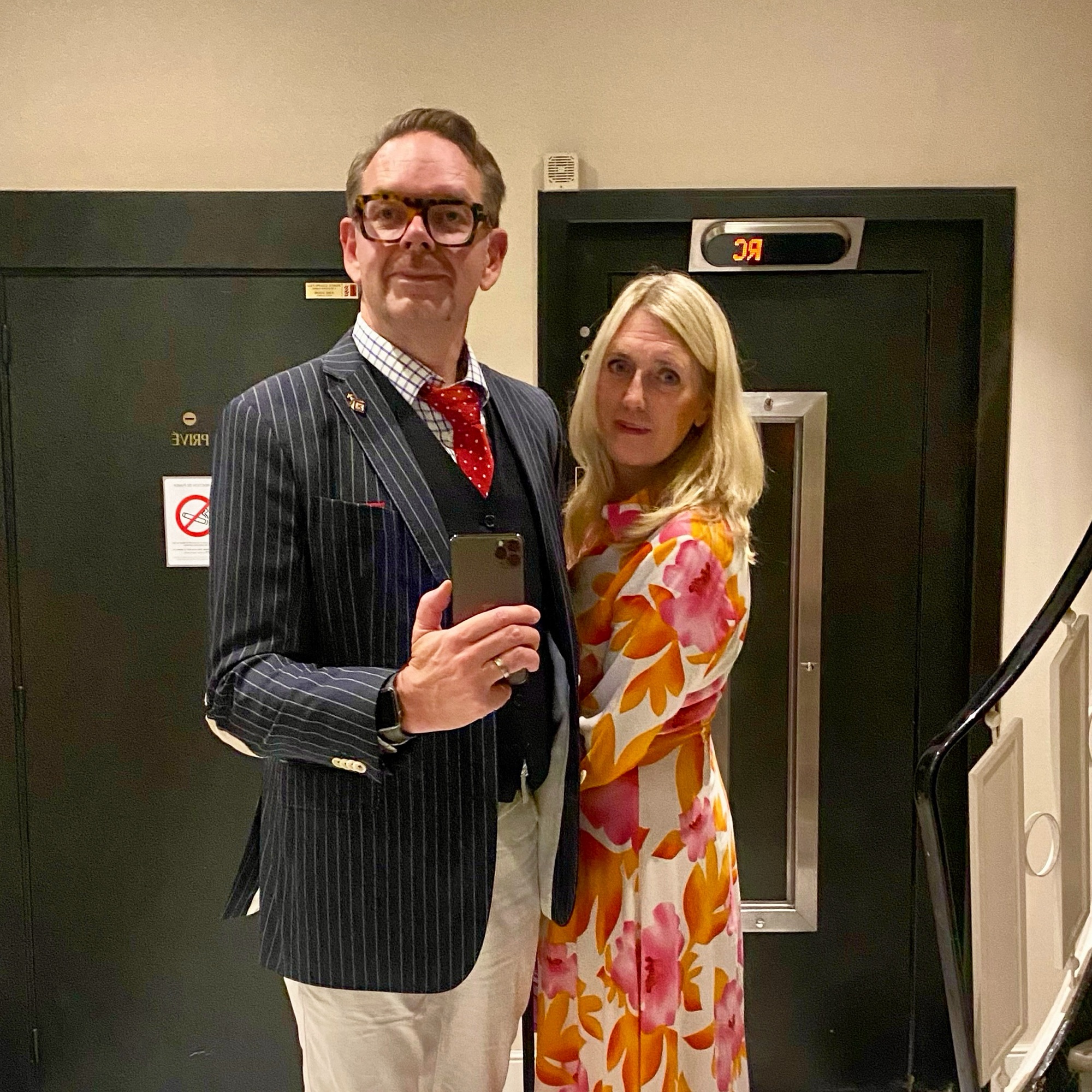 A man in a pinstripe suit and a woman in a floral dress posing together in a mirror.