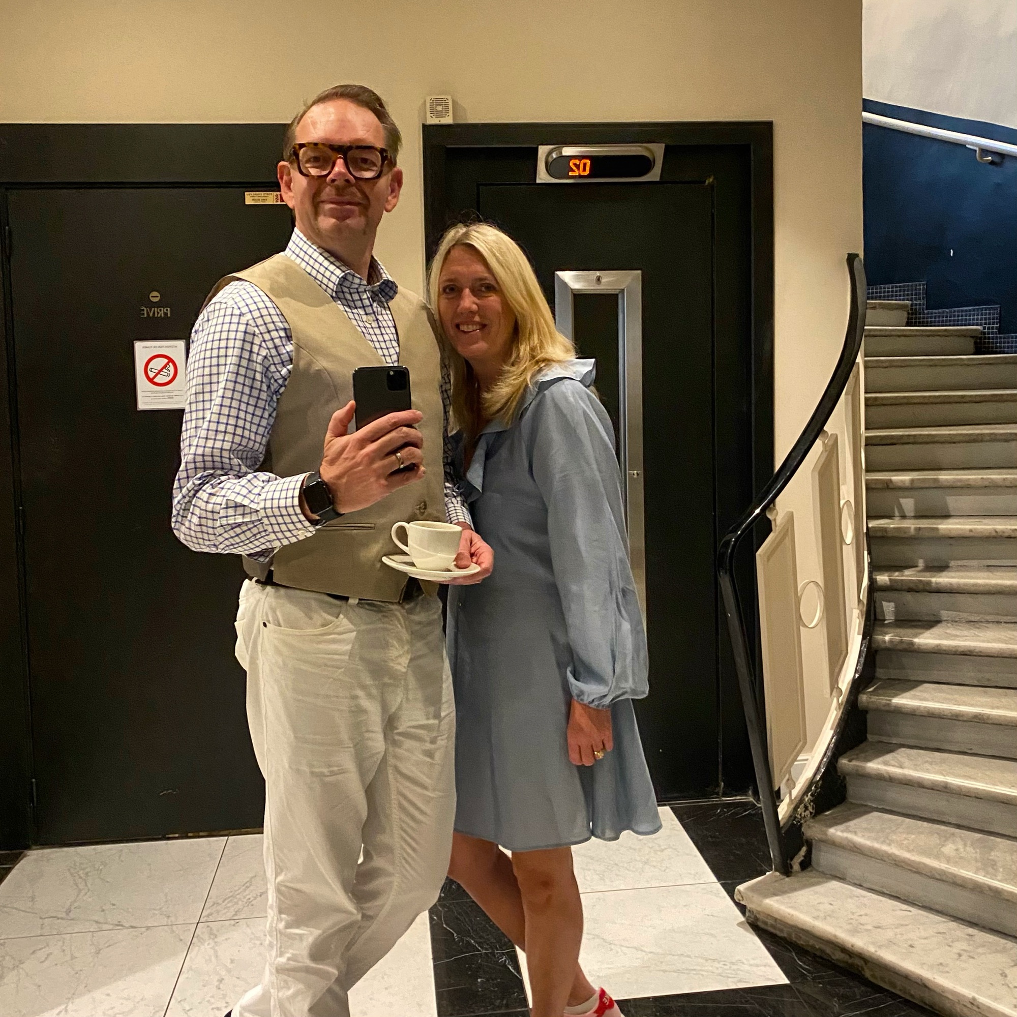 A man in glasses wearing a beige vest and white trousers, holding a cup, stands next to a woman in a light blue dress.