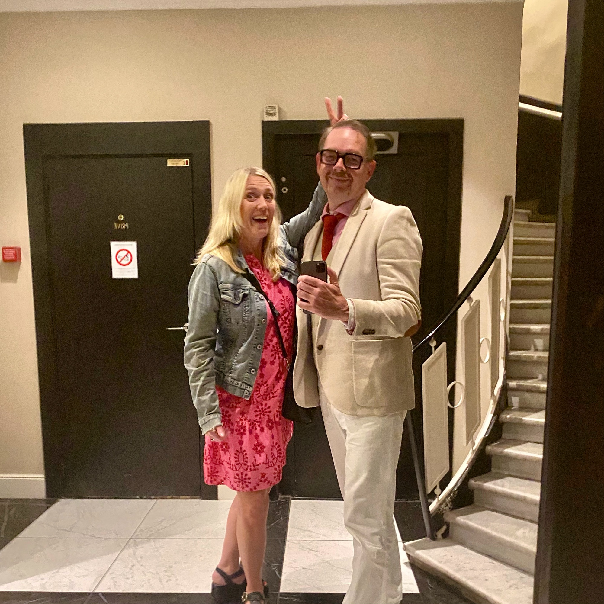 A man in a white suit and glasses, and a woman in a pink dress with a denim jacket, posing in a mirror.