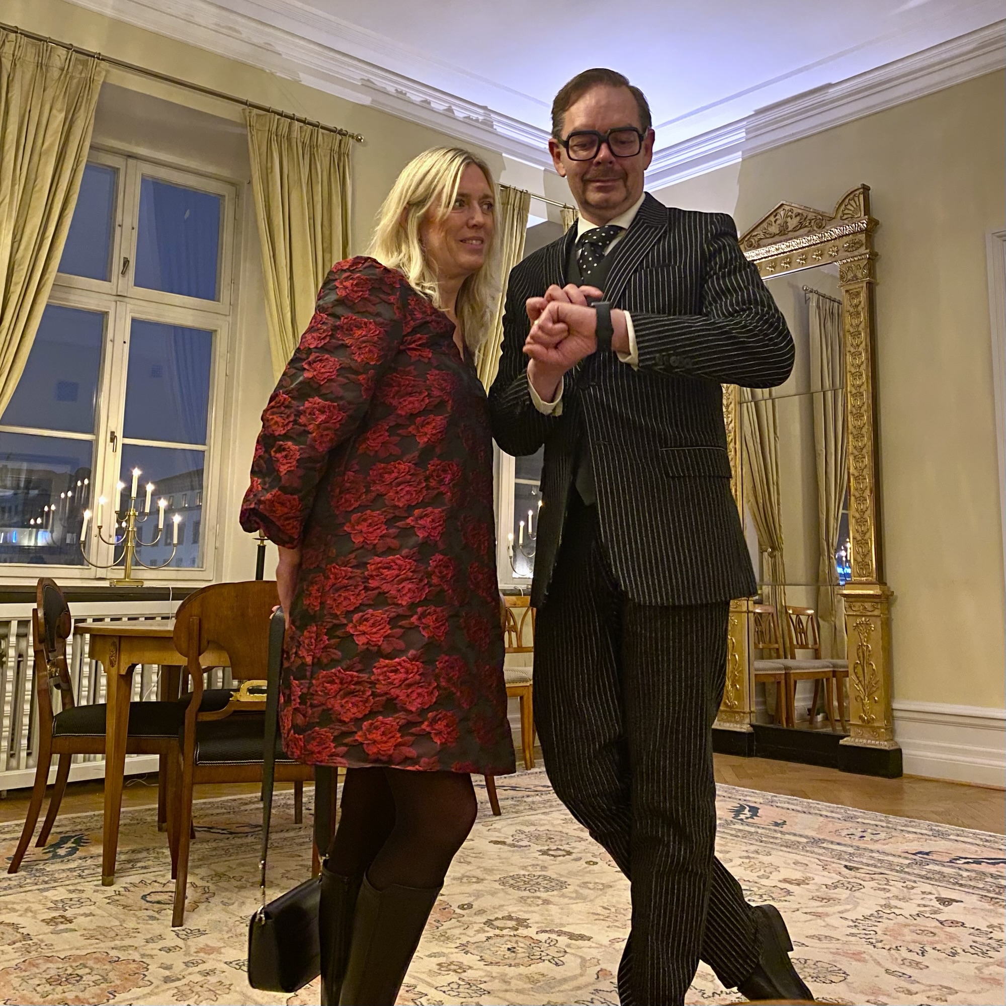 A man in a pinstripe suit and glasses stands next to a woman in a floral coat, both smiling in an elegant indoor setting.