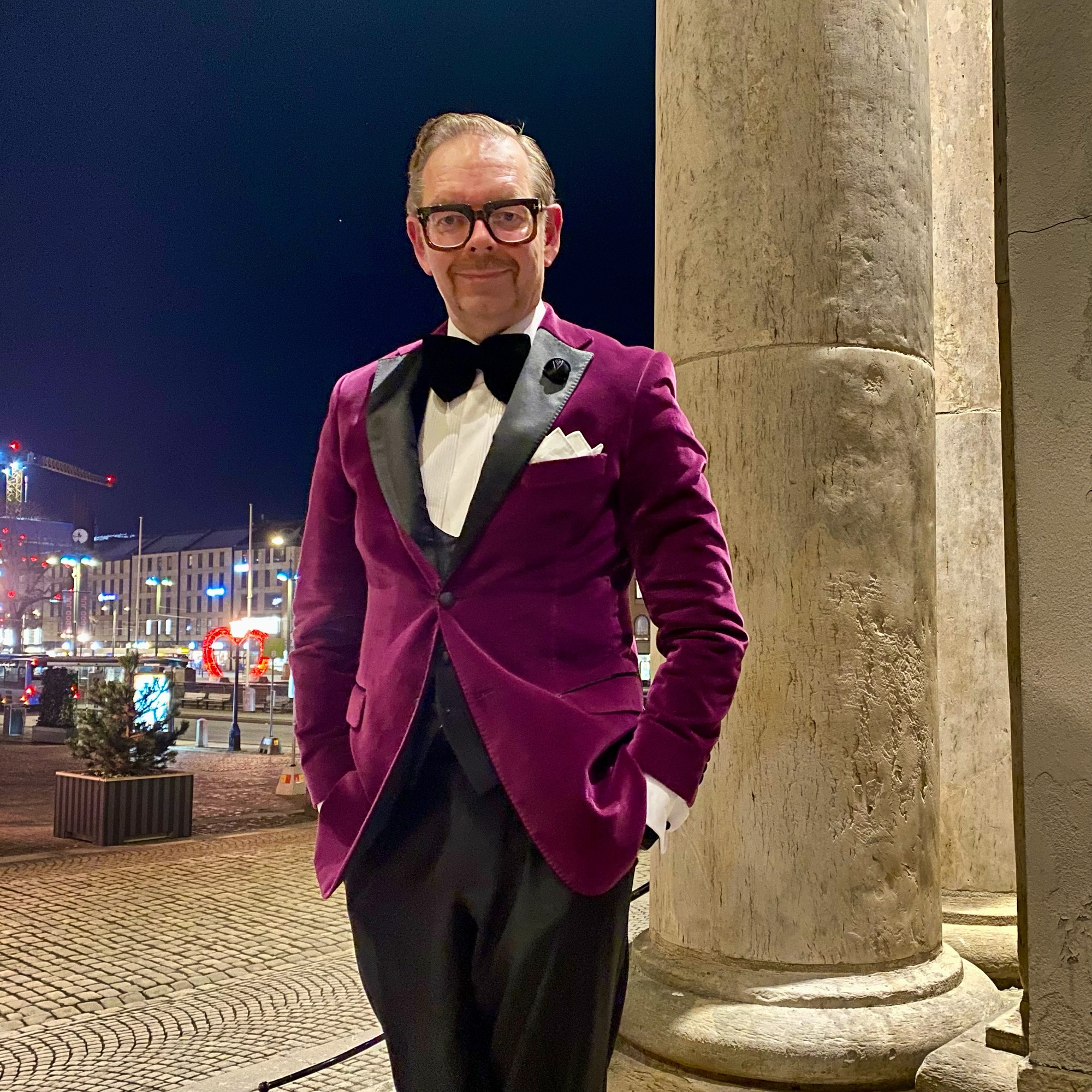 Man wearing a burgundy velvet tuxedo with a black bow tie, standing confidently near a column at night.