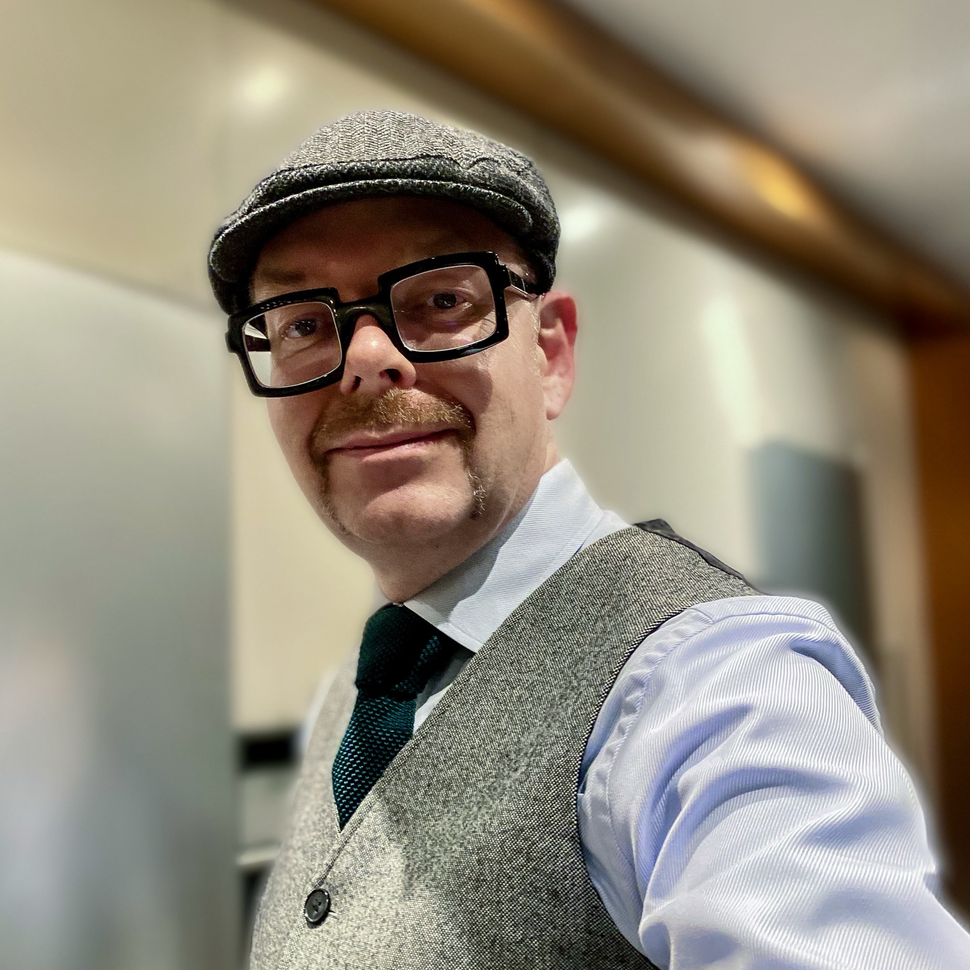 Man wearing a grey flat cap, glasses, and a waistcoat, posing for a self-portrait indoors.
