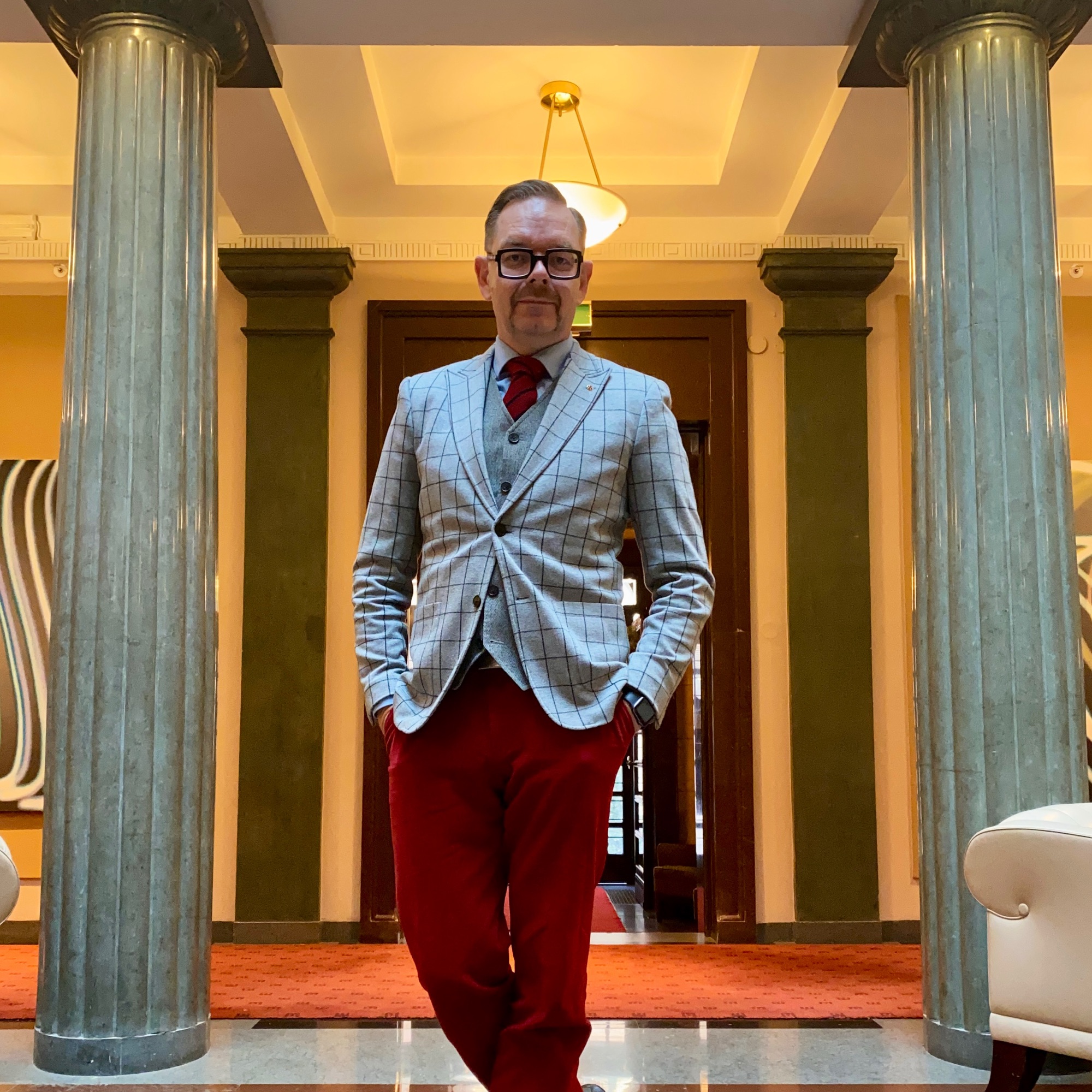 Man in a grey checked blazer and red trousers standing in a grand interior with columns.