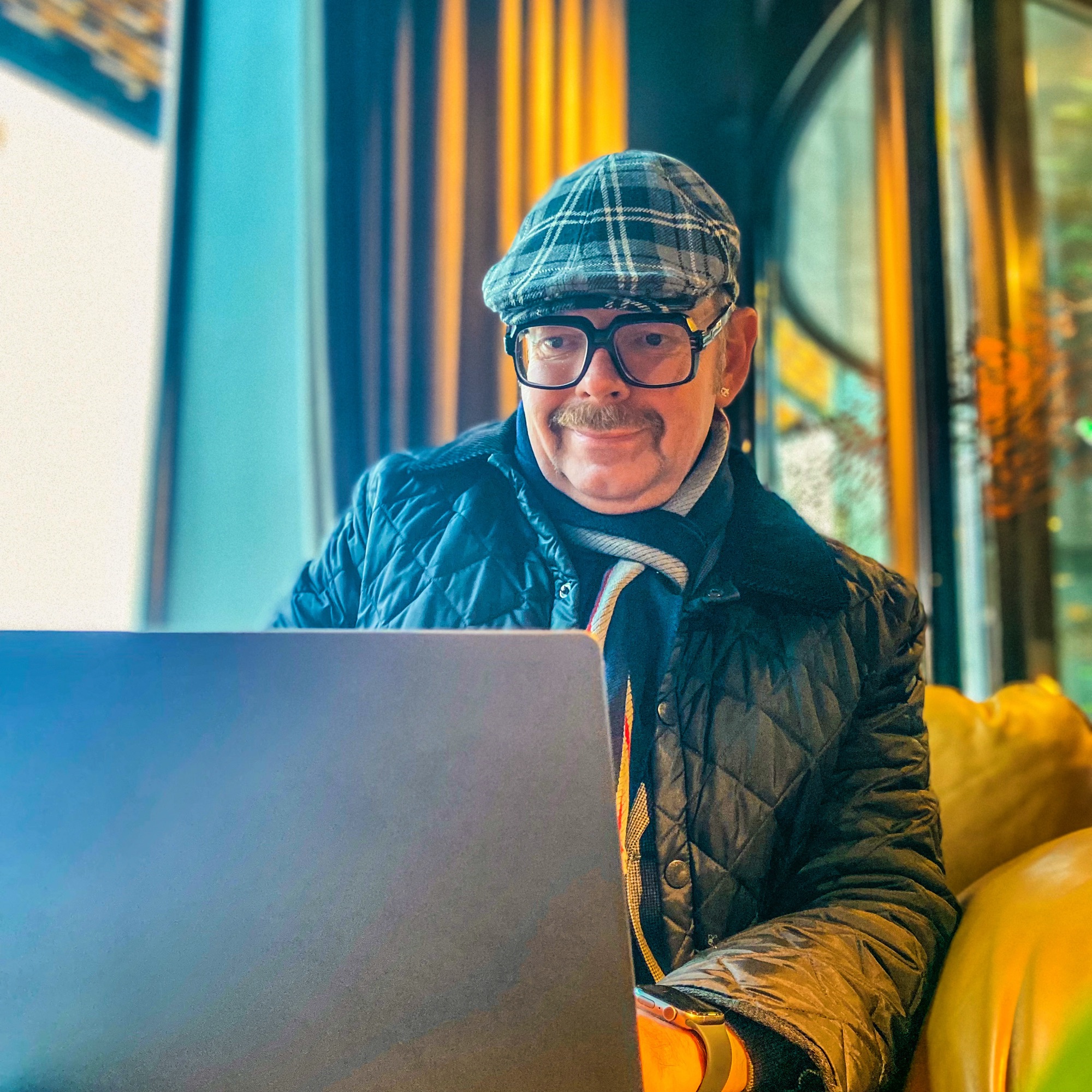 Man wearing a flat cap and glasses, seated with a laptop in a well-lit indoor space.