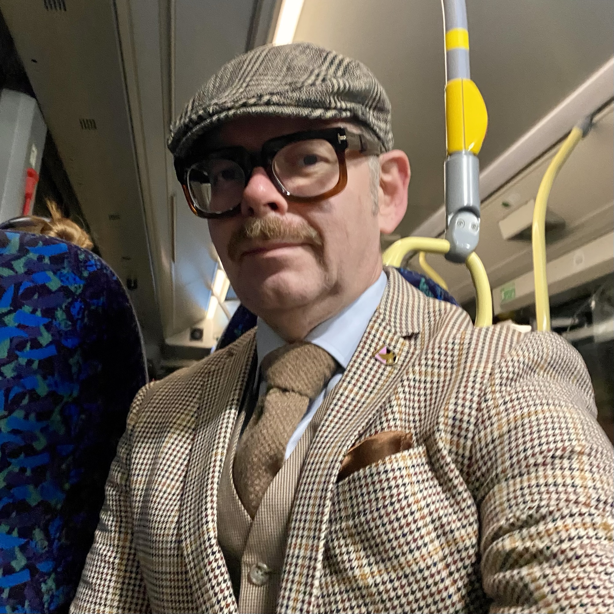 Man wearing a patterned suit and flat cap, seated on a bus with a serious expression.