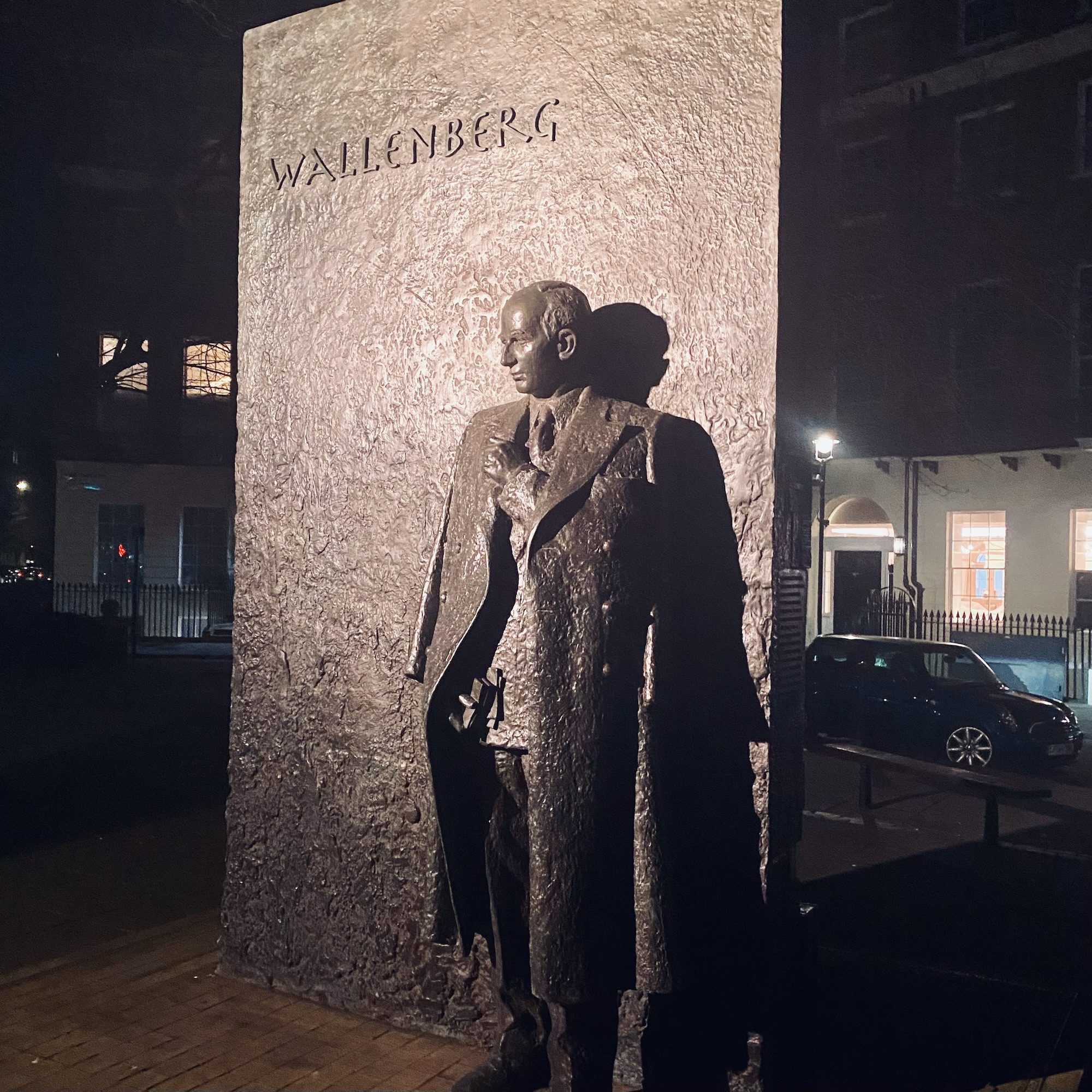 Bronze statue of Raoul Wallenberg standing beside a textured stone wall with his name inscribed.