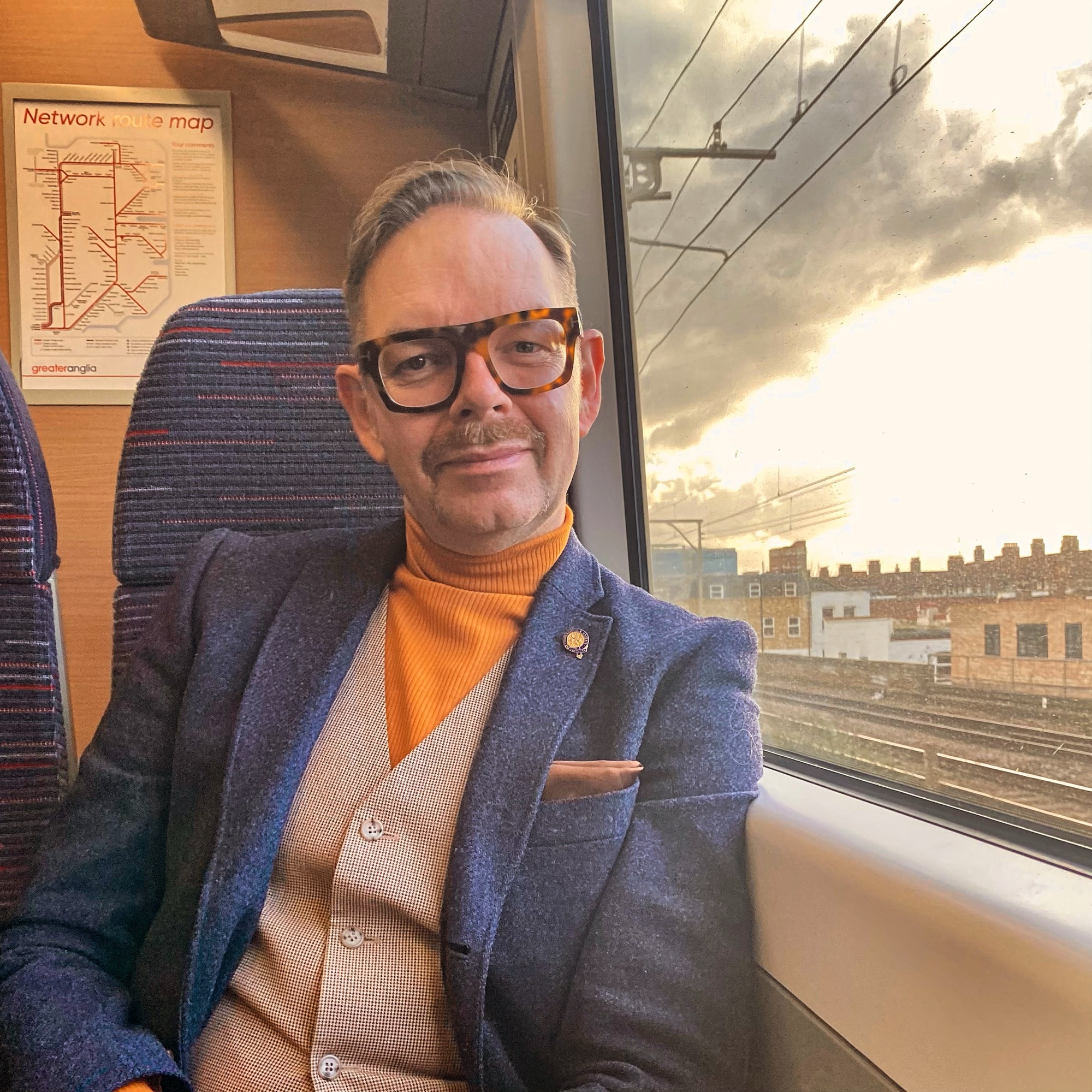 Man with glasses and a moustache sitting on a train, wearing a turtleneck and blazer, with a train map in the background.