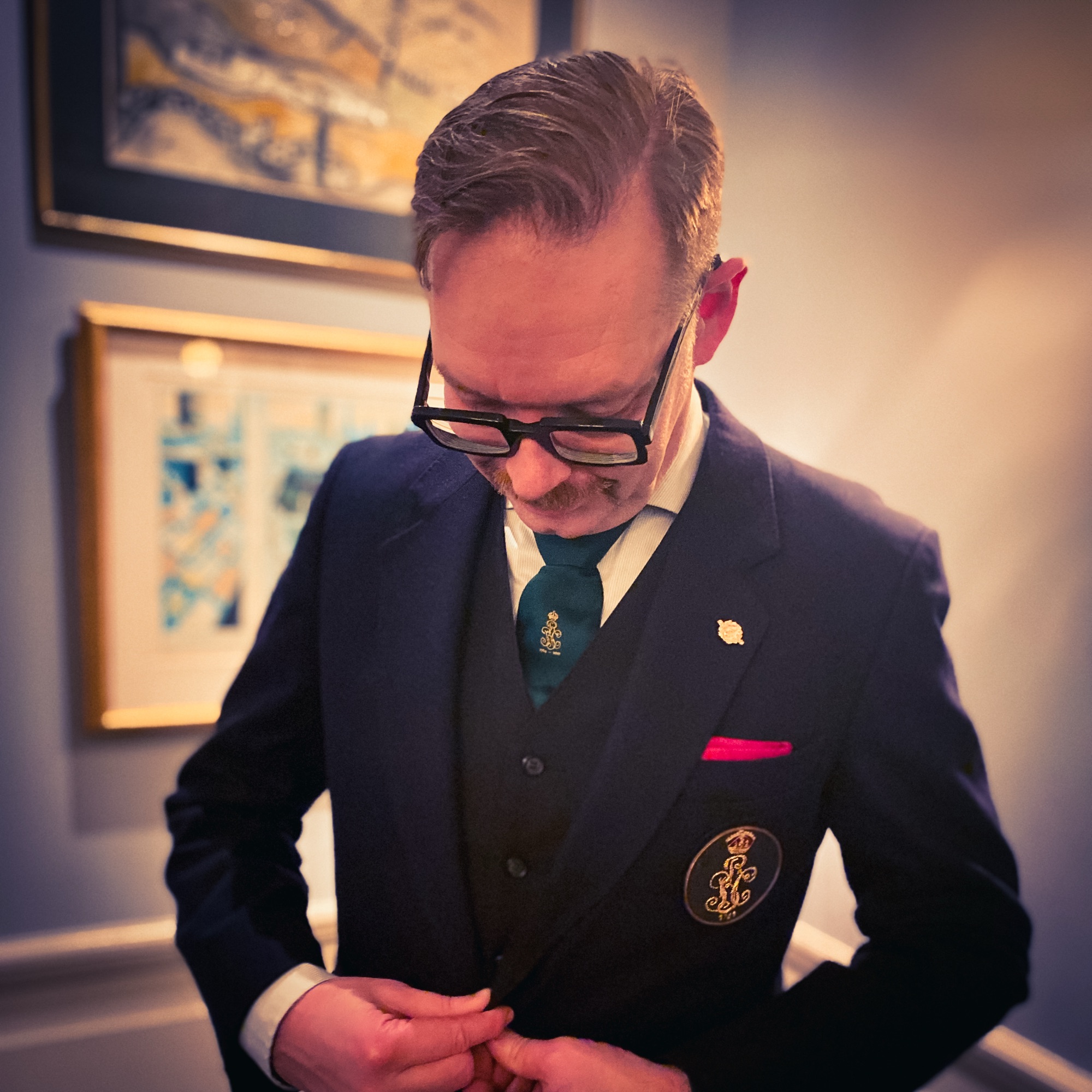 Man adjusting the buttons on a dark suit jacket while wearing glasses and a tie with a small emblem.