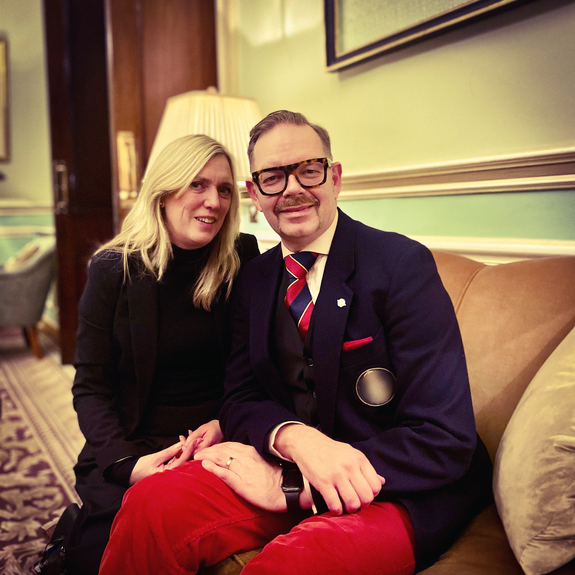 A man in a suit with glasses and a woman in a black outfit sitting together on a sofa in a formal setting.