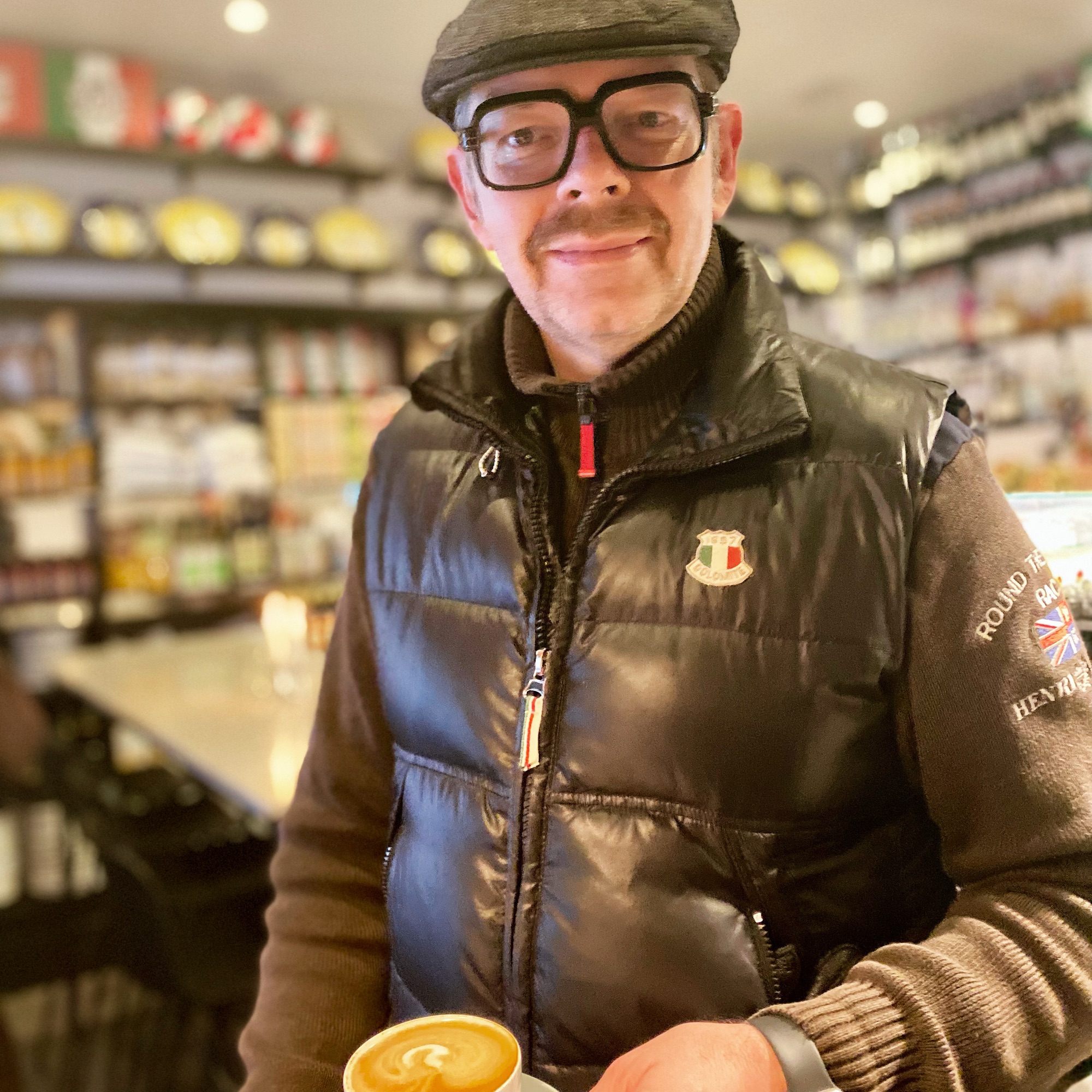 Man in a black puffer vest and glasses holding a cup of coffee in a café setting.