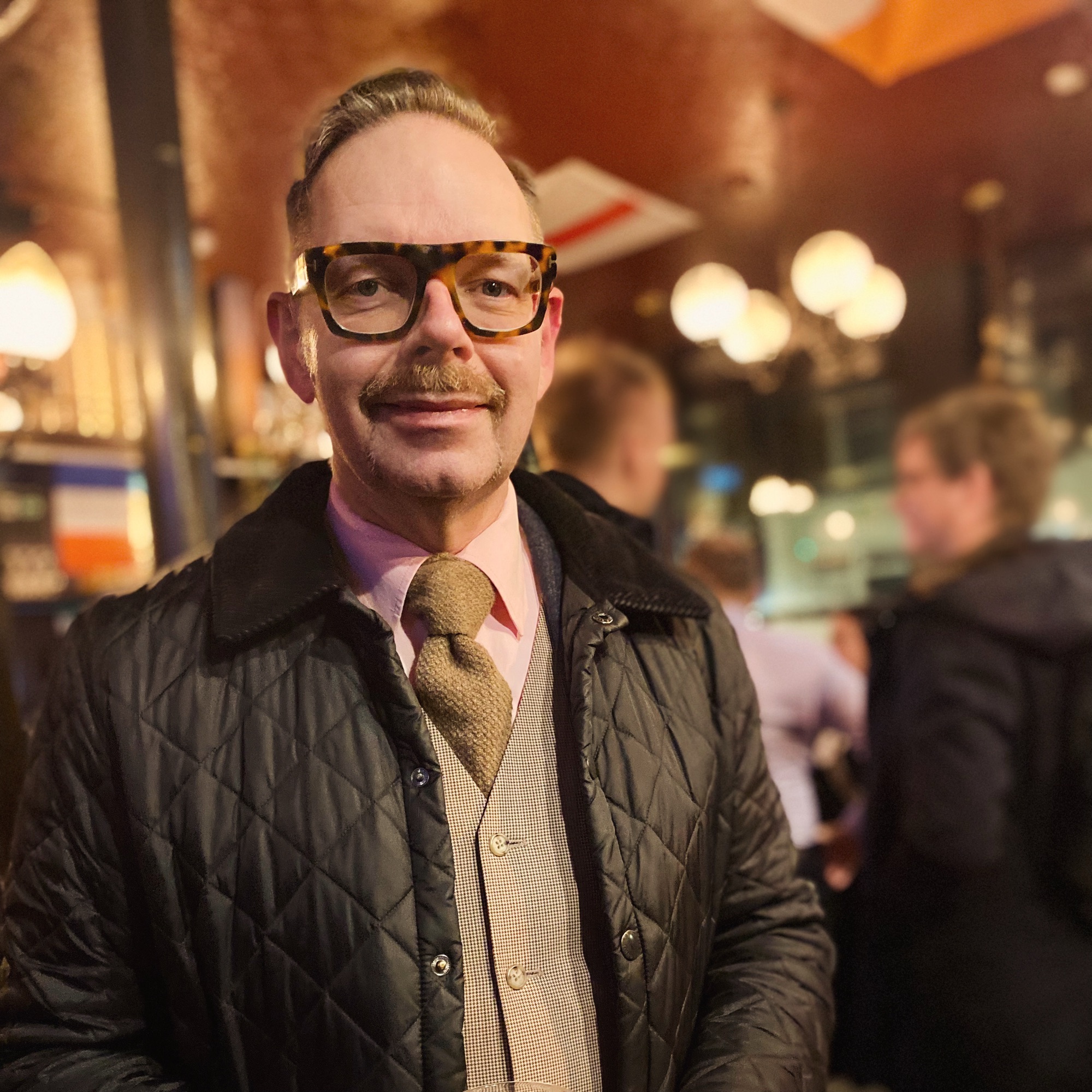 Man wearing glasses and a patterned tie, dressed in a quilted jacket and vest, standing indoors.
