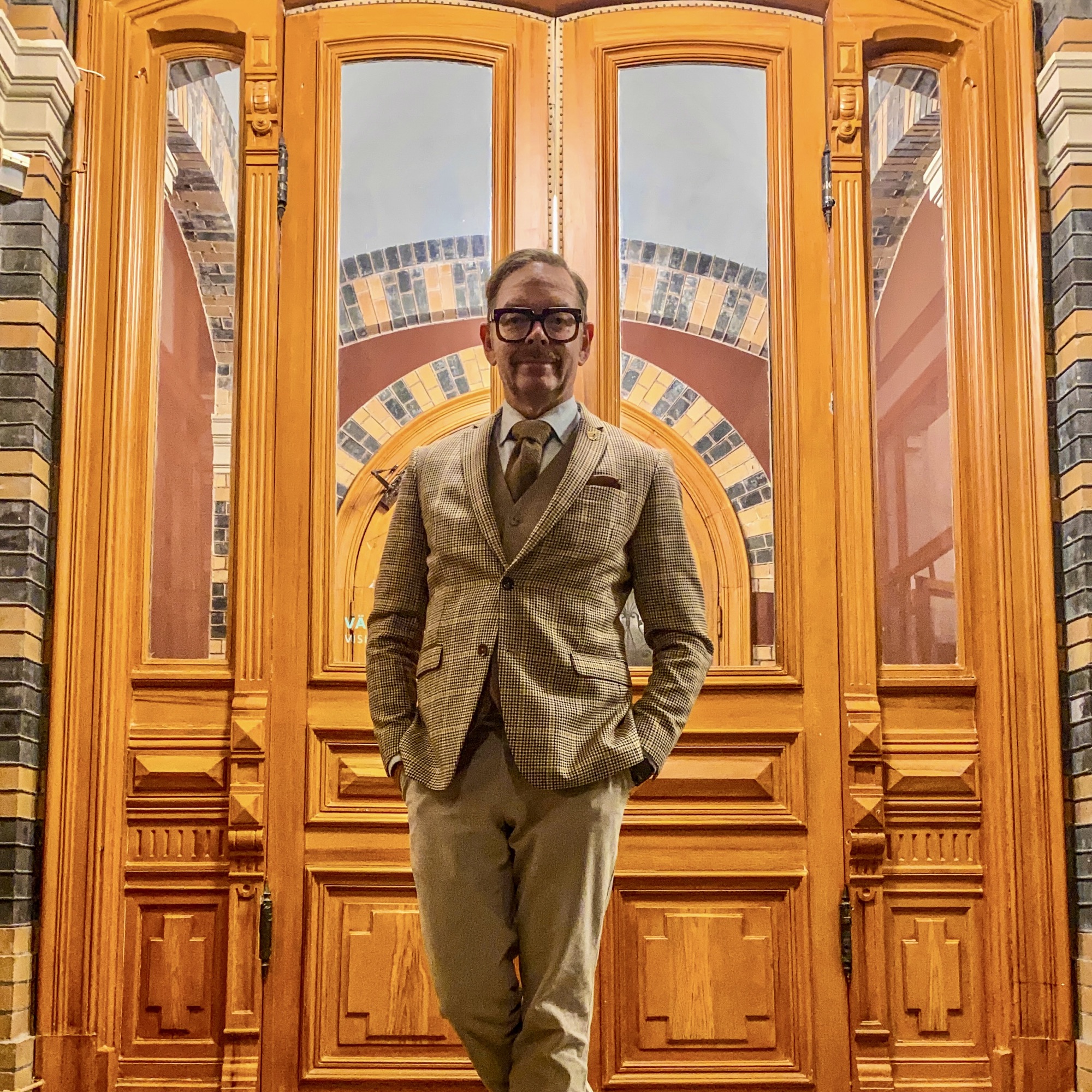 Man in a checked suit standing confidently in front of a large wooden door with intricate designs.