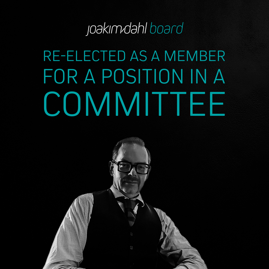 A man in formal attire with glasses, seated and looking at the camera, with text stating 'Re-elected as a member for a position in a committee'.