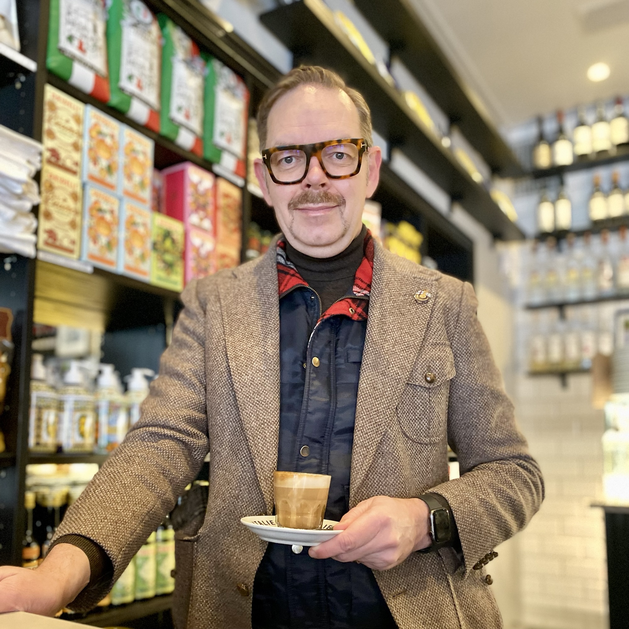 Man in a brown blazer holding a coffee cup on a saucer in a café with shelves of products behind him.