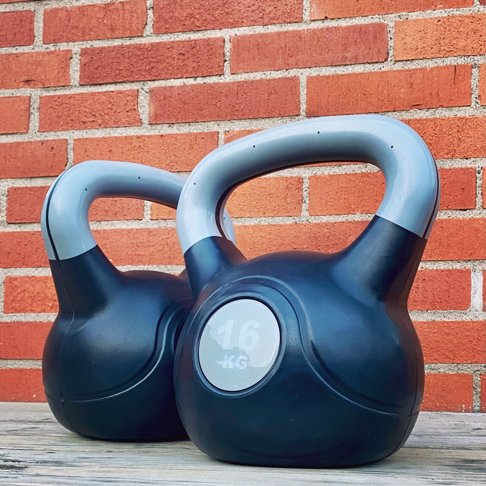 Two black kettlebells with grey handles, each weighing 16 kg, positioned against a brick wall.