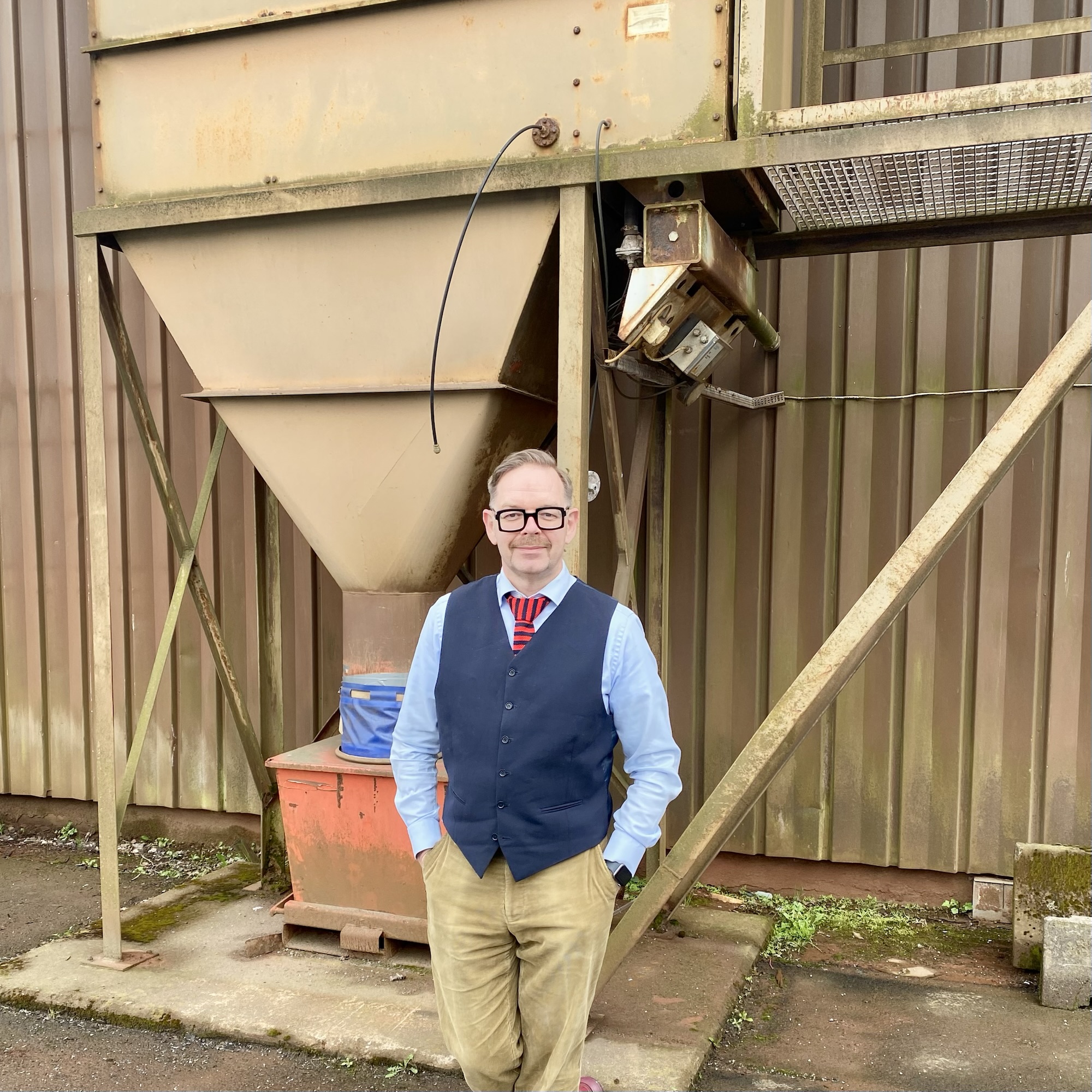 Man in a blue shirt and grey vest standing beside an industrial hopper in a manufacturing setting.