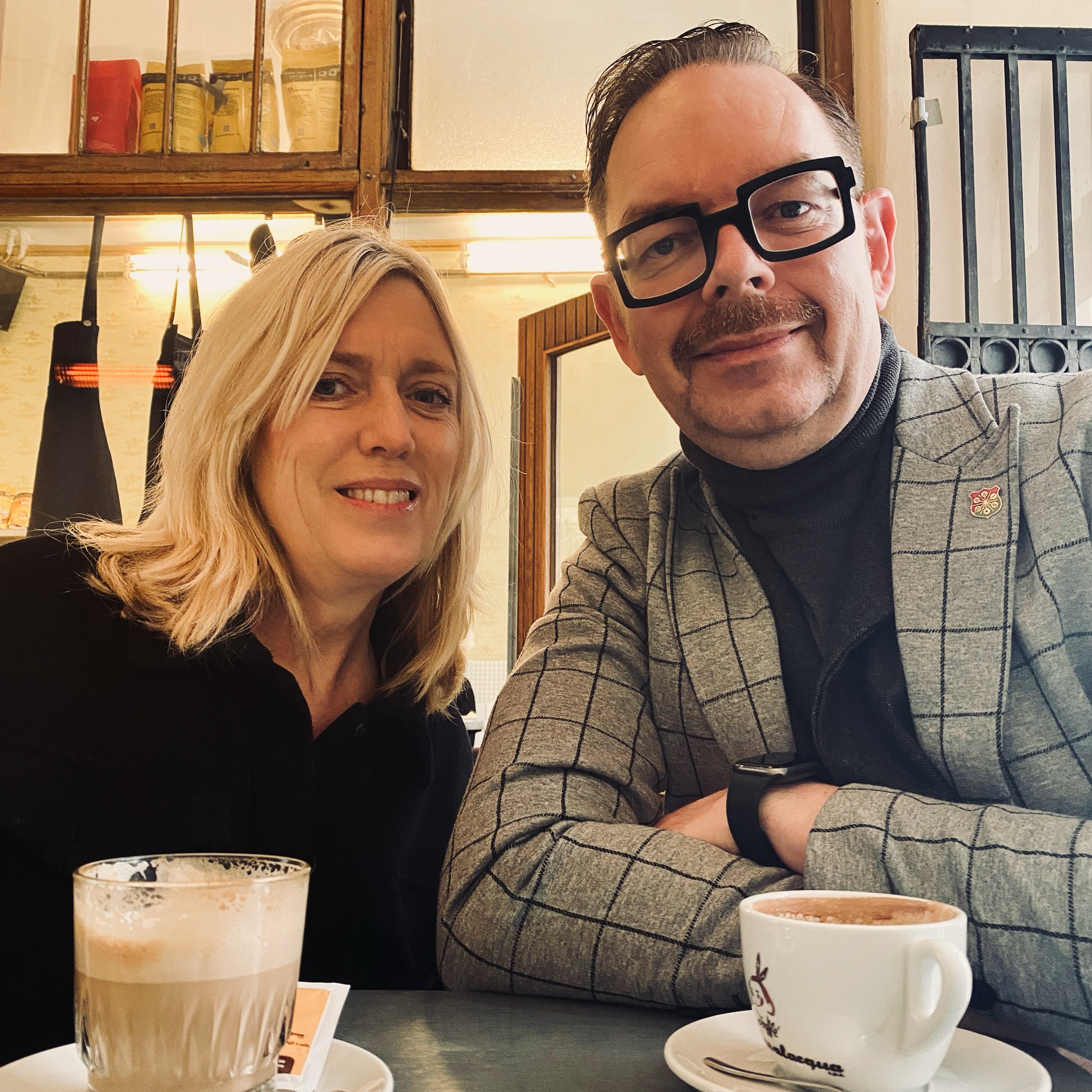 Two individuals seated at a table with coffee cups, one wearing glasses and a grey blazer, the other with blonde hair.