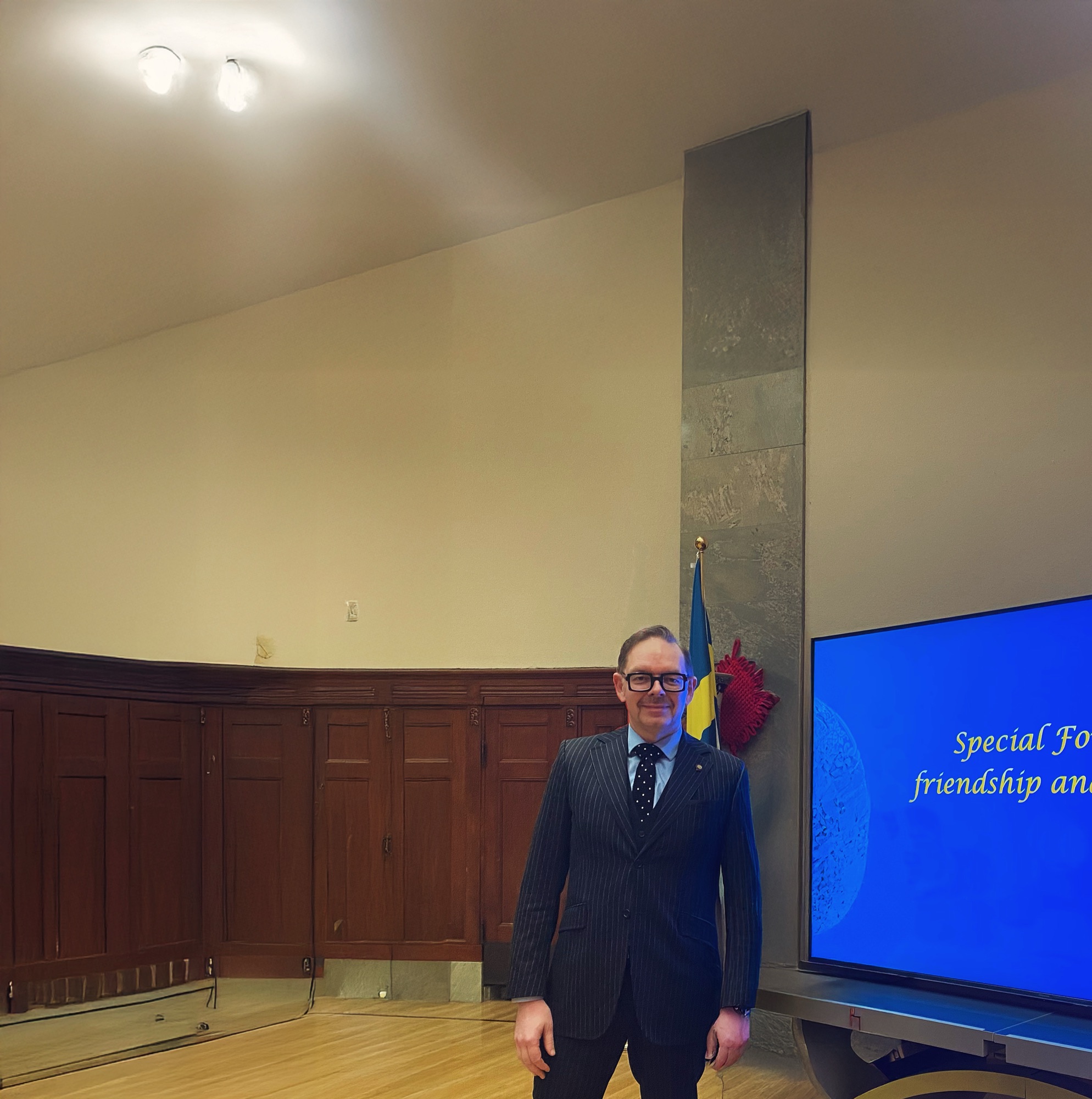 Man in a pinstripe suit standing in a room with wooden paneling and a screen displaying text about friendship and cooperation.