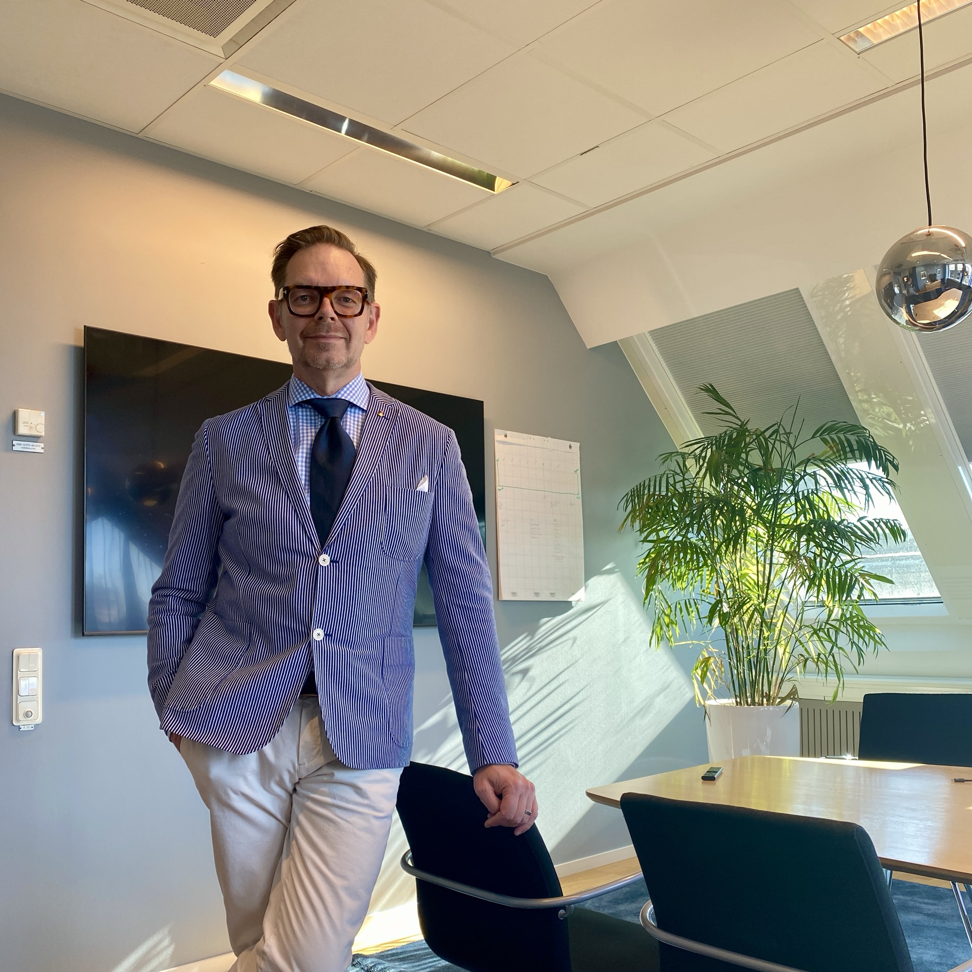 Man in a blue checked blazer and white trousers standing in a modern office with a plant and large screen.
