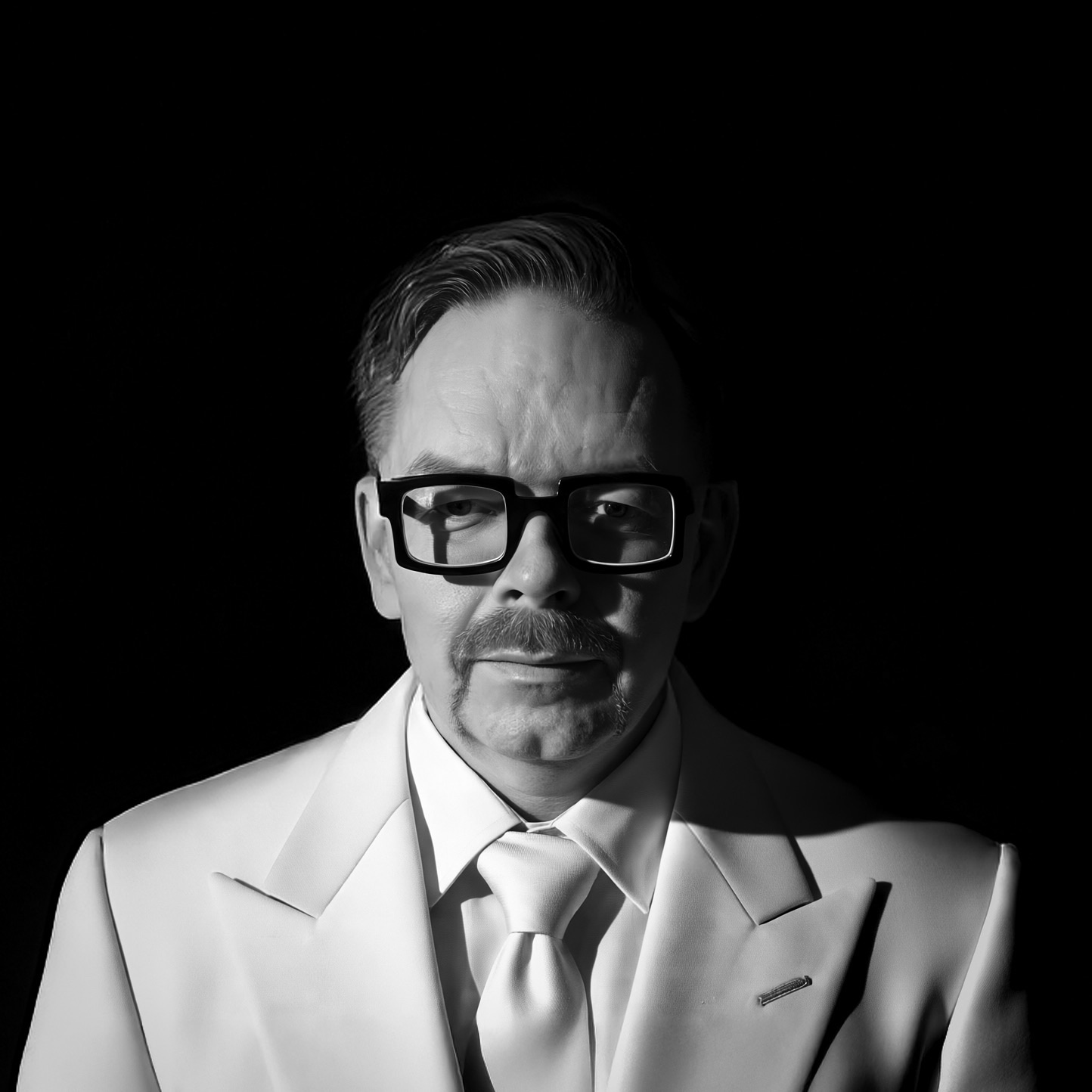 Man in a white suit and glasses with a moustache, set against a dark background.