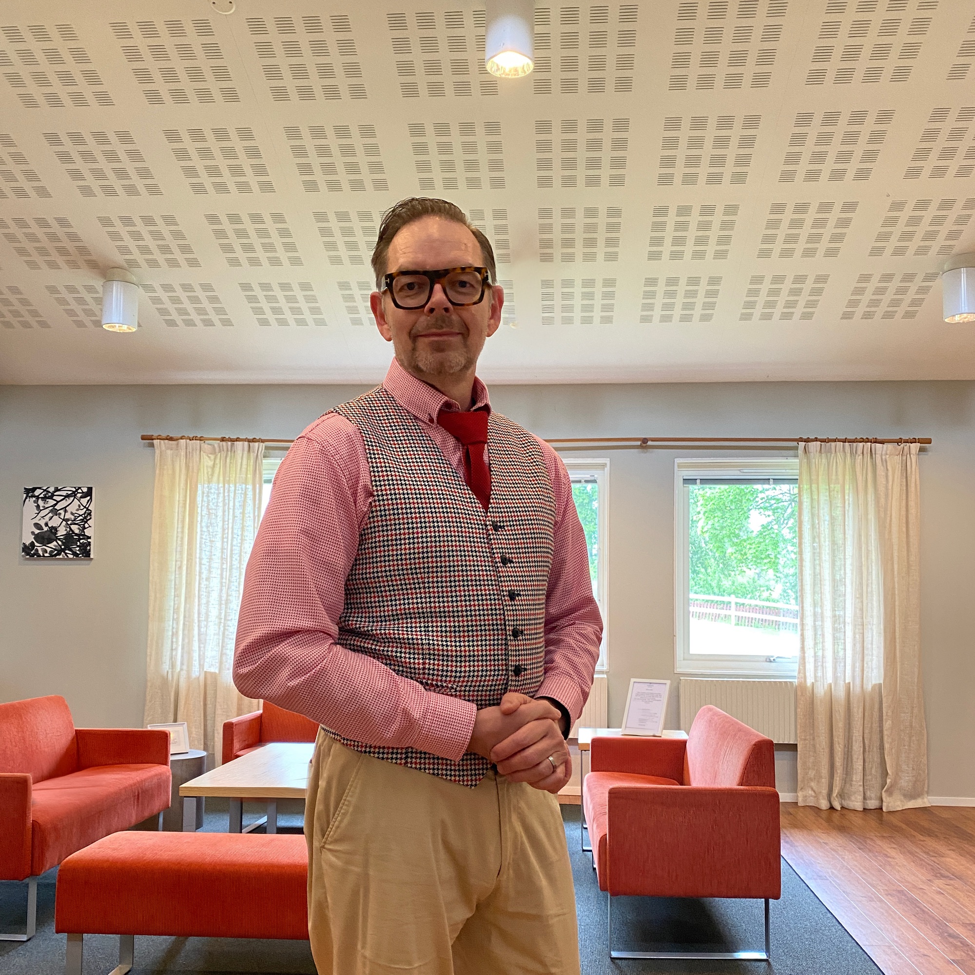 Man wearing a checked vest and red tie, standing in a room with orange furniture and large windows.
