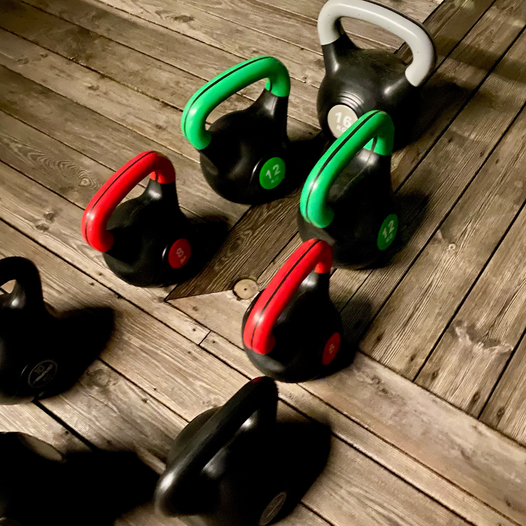 A collection of kettlebells in various weights, including red, green, and grey handles, arranged on a wooden floor.