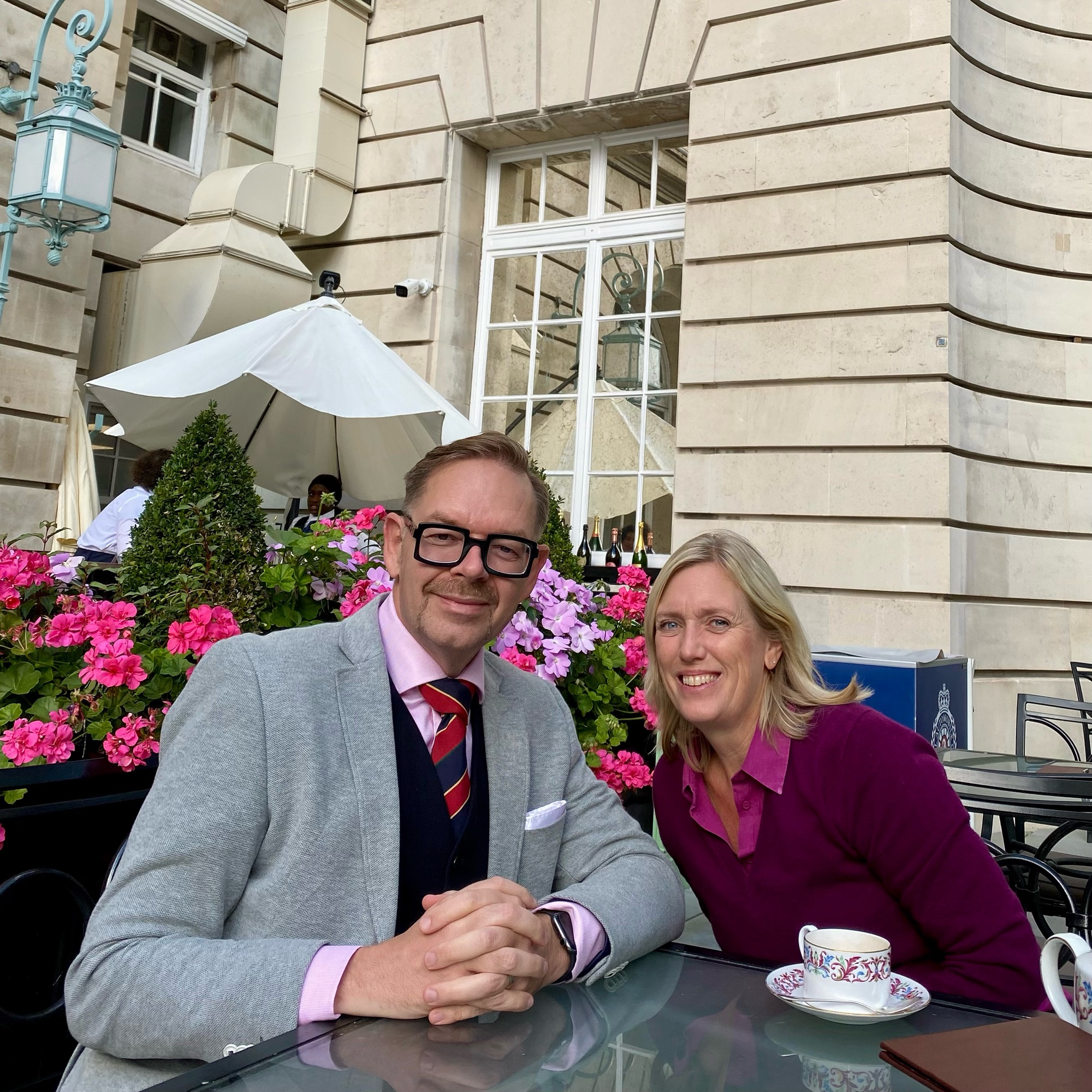 Two individuals seated at a table with a cup of tea, surrounded by pink flowers at a club on Pall Mall.