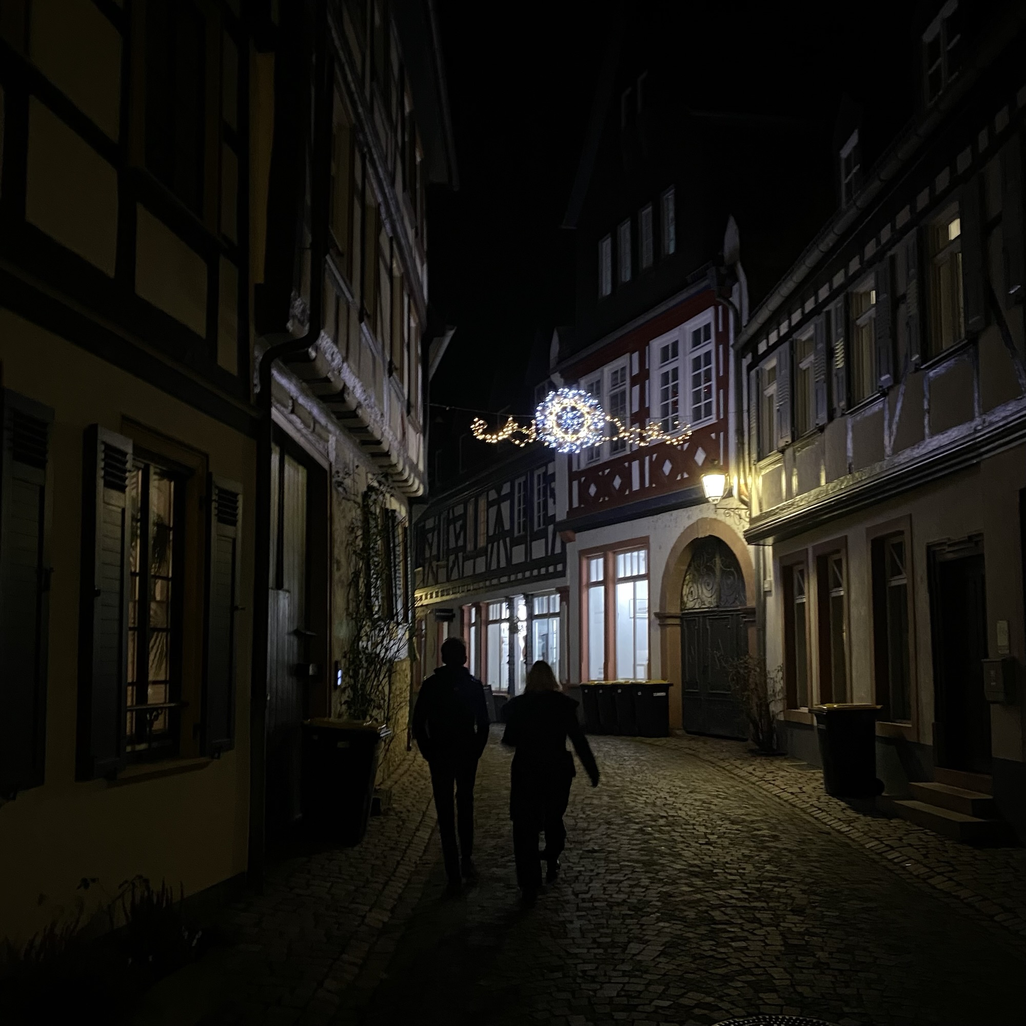 Two people walking down a cobblestone street at night, illuminated by streetlights and festive decorations.