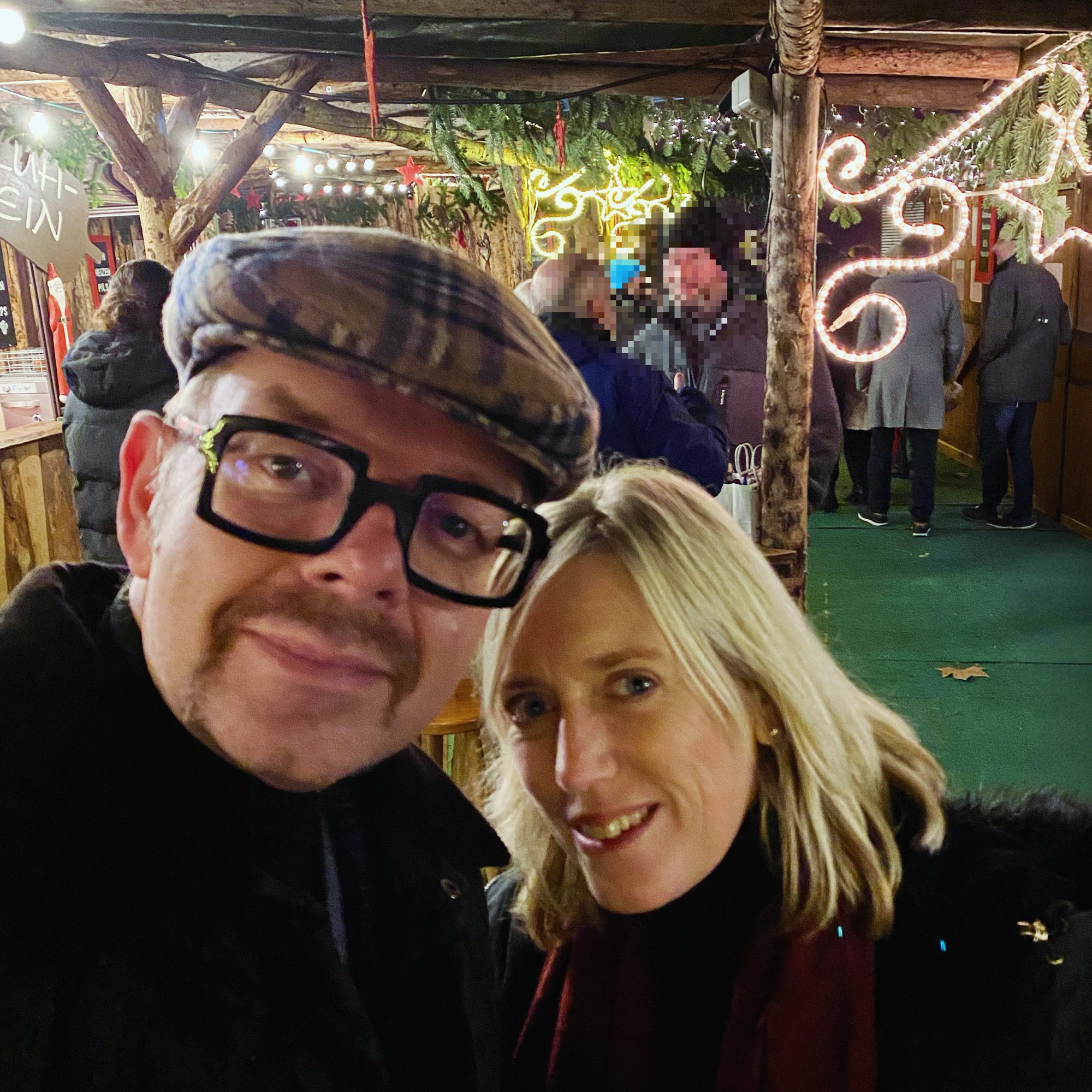 Man wearing a flat cap and glasses posing with a woman in a festive setting with lights in the background.