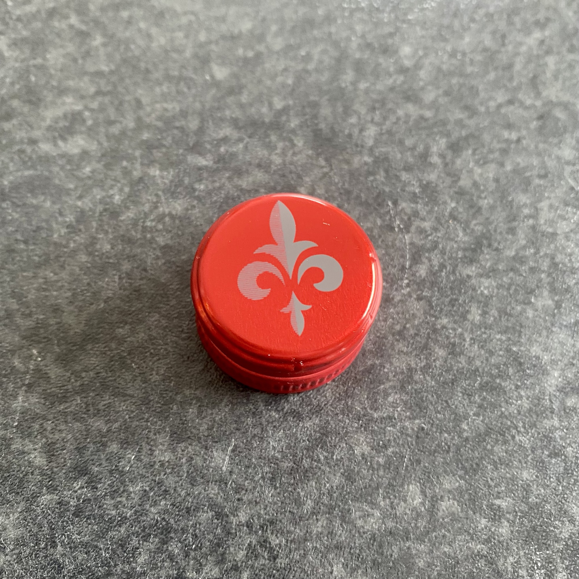Red bottle cap with a silver fleur-de-lis design on a textured surface.
