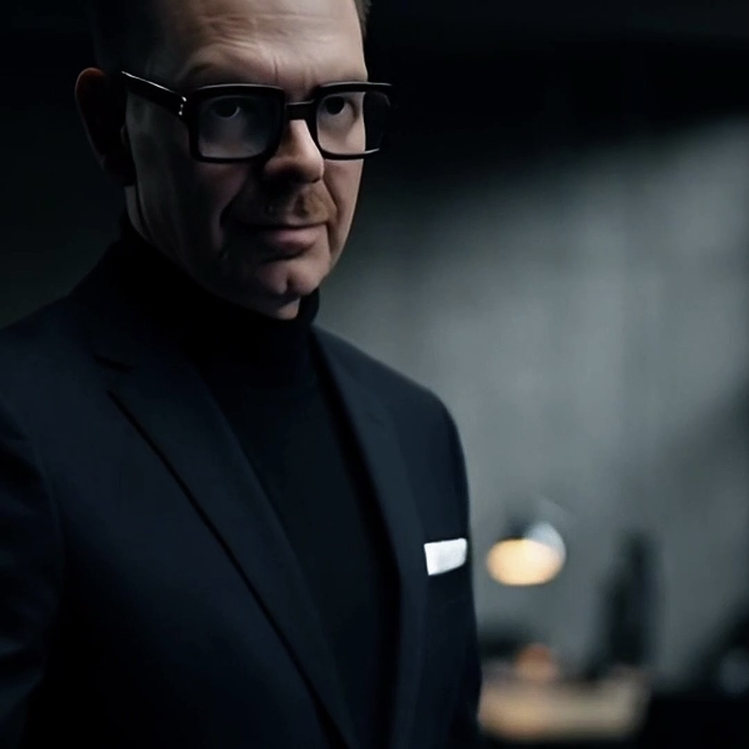 Man in a black suit and glasses, looking directly at the camera with a serious expression in a dimly lit environment.
