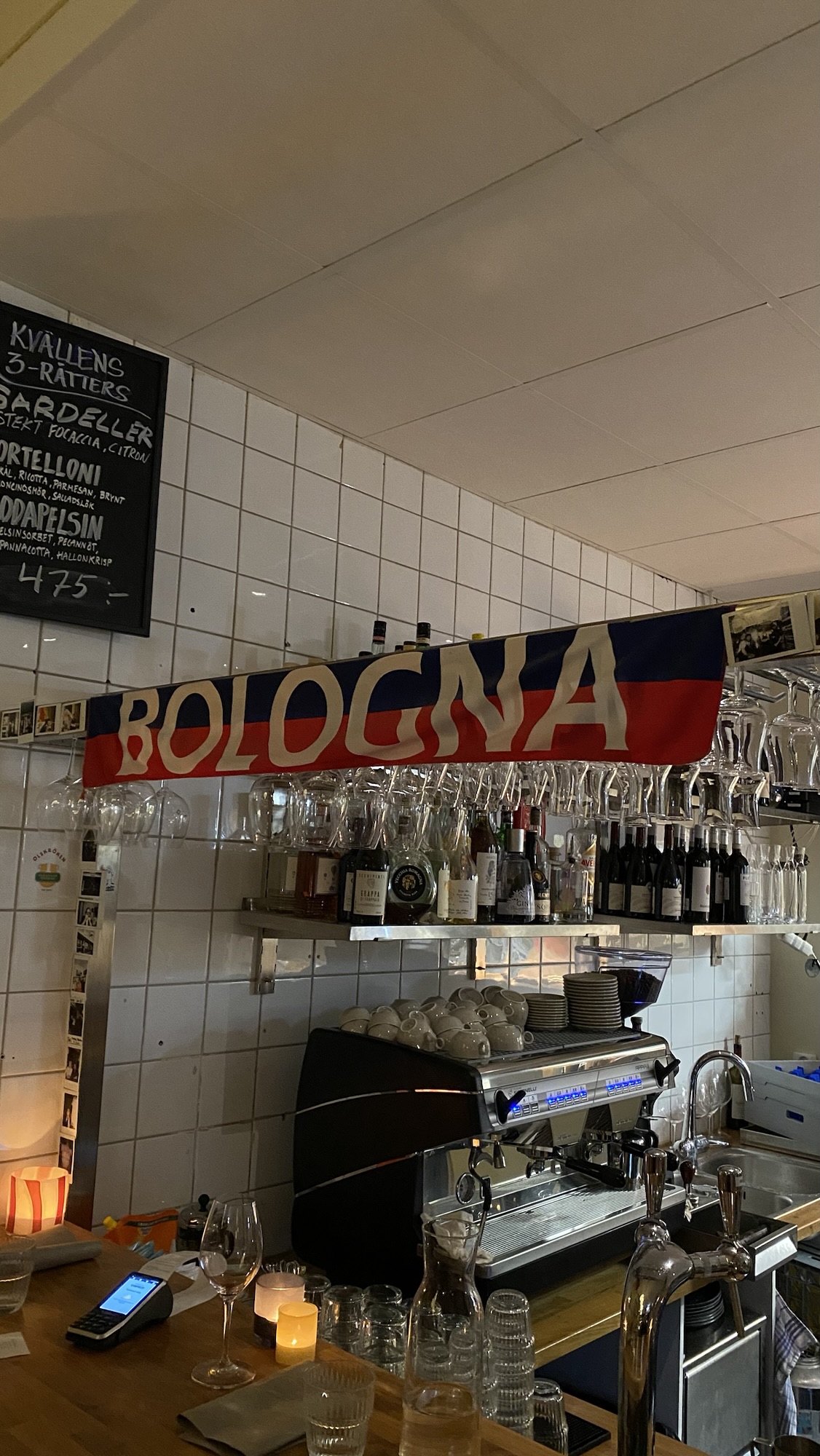 Bologna football team scarf in dark blue and red stripes above a coffee counter, with rows of glasses and bottles in an Italian street restaurant.
