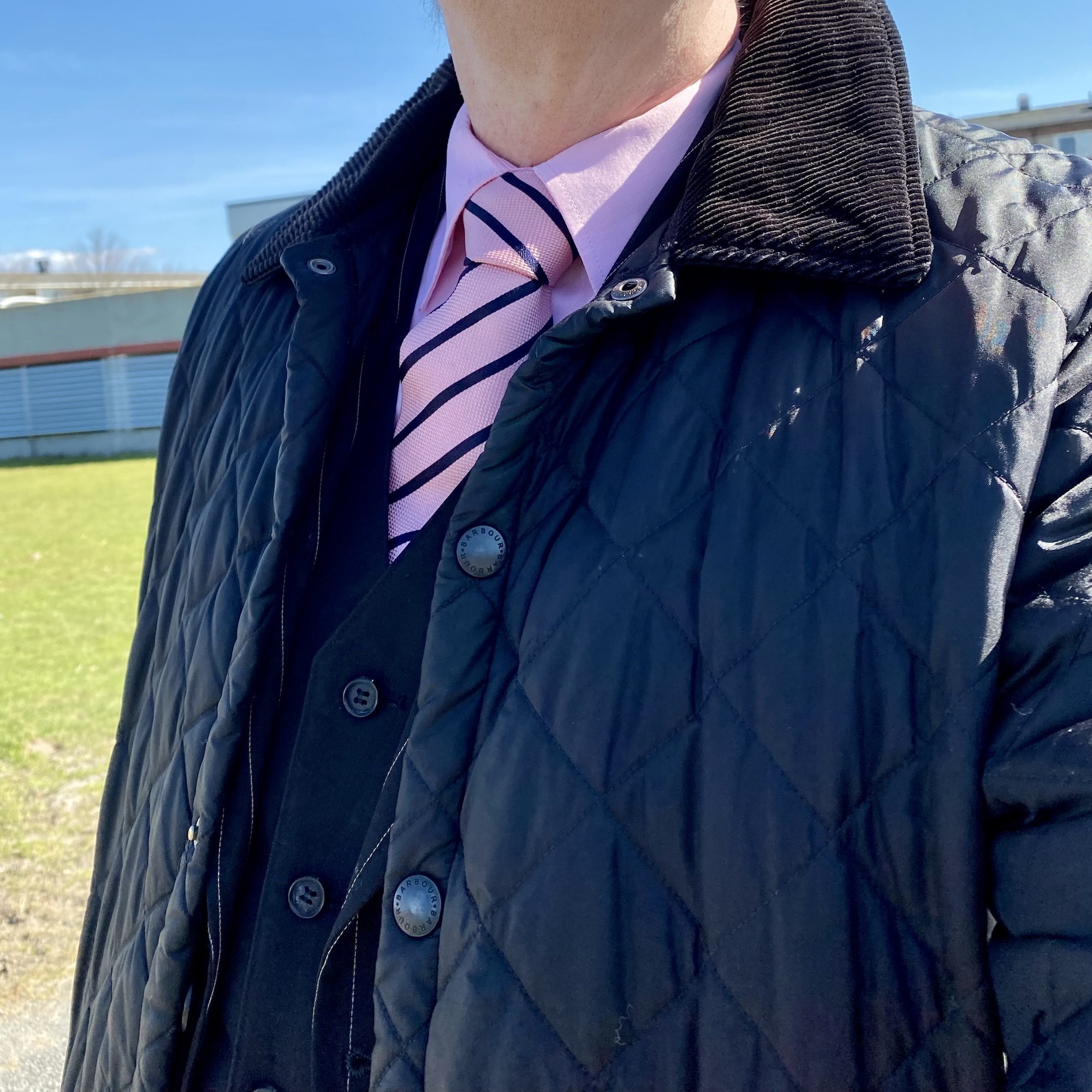 Joakim Dahl-inspired look with a pink shirt and striped tie beneath a navy quilted Barbour jacket, complemented by a Tiger of Sweden suit, photographed outdoors.
