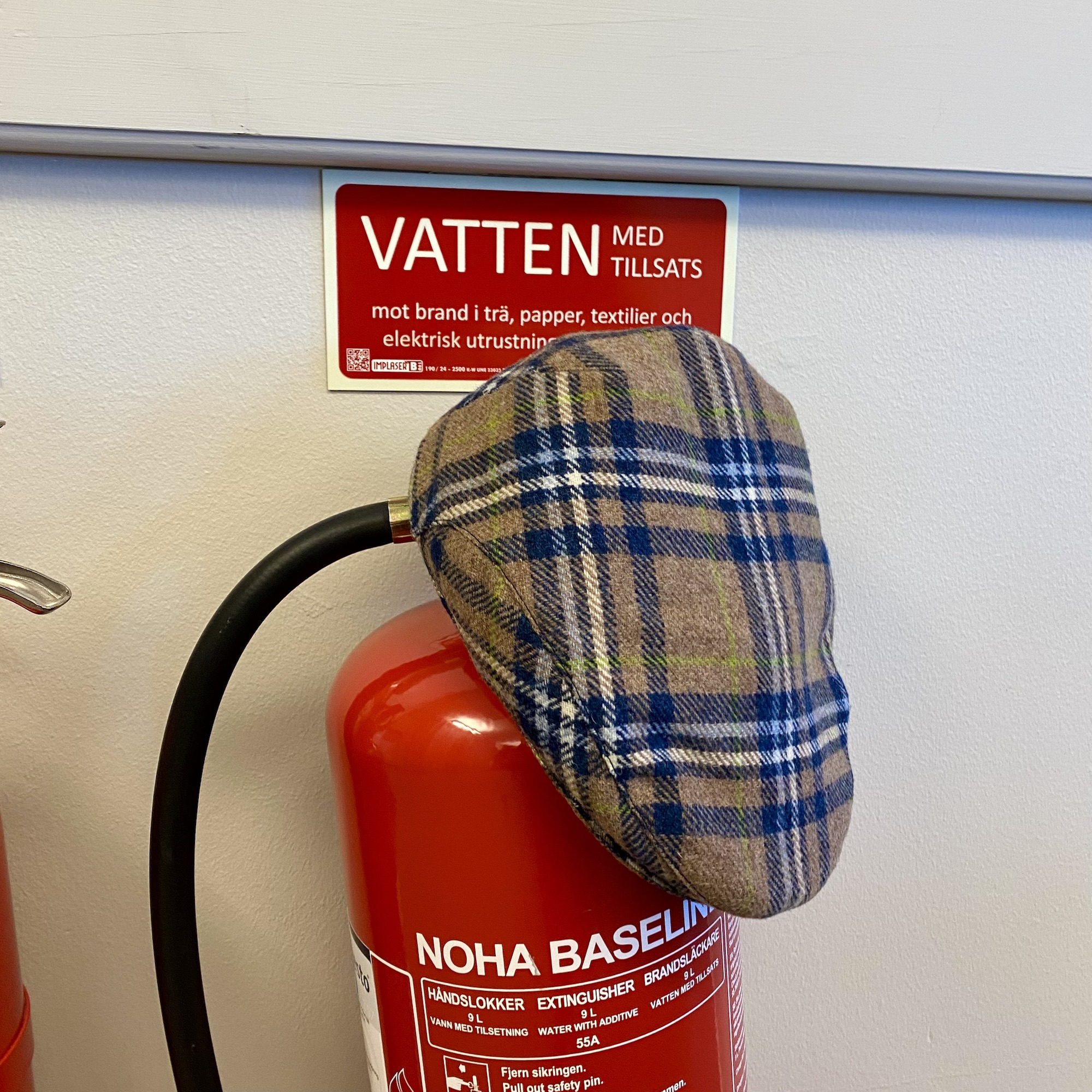 Blue and grey check flat cap resting on a red NOHA BASELINE fire extinguisher, with “VATTEN MED TILLSATS” visible on the label.