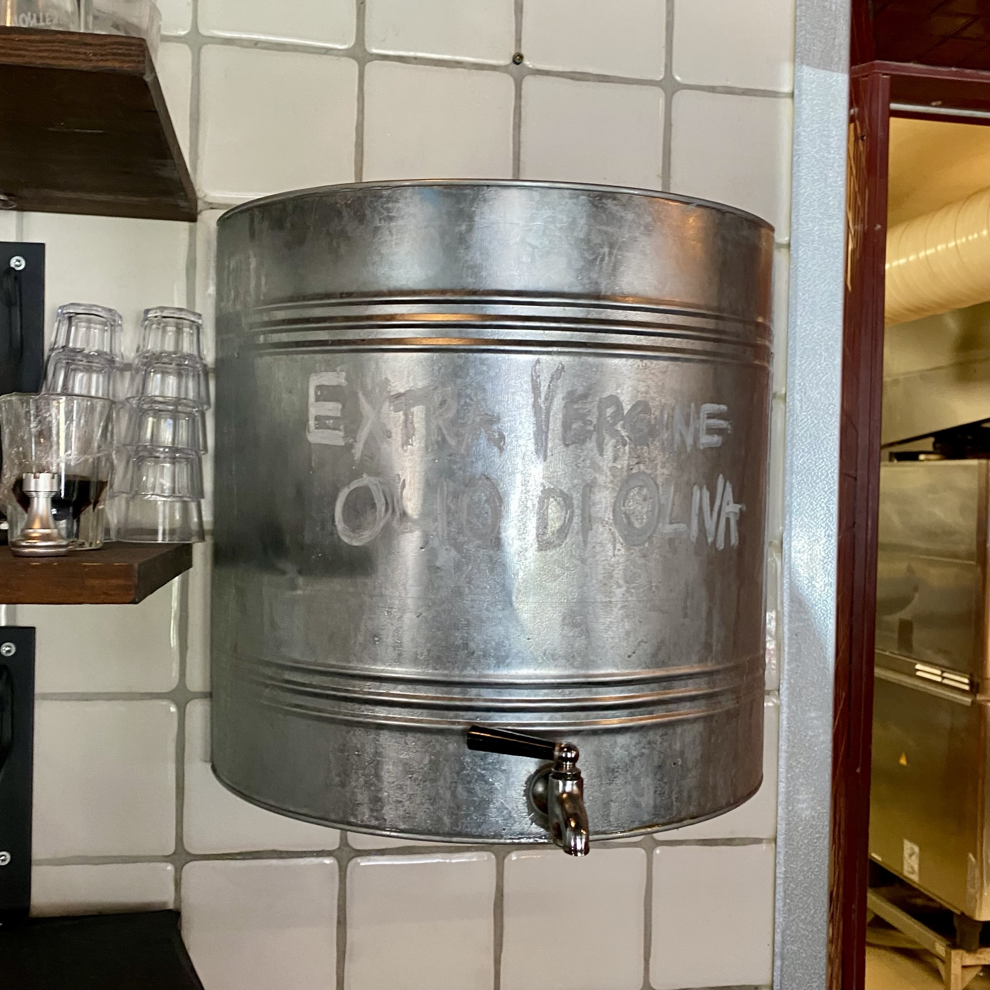 Metallic cylindrical dispenser with a spigot, embossed weathered lettering, mounted on a tiled wall; evokes a rustic olive oil serving scene linked to Joakim Dahl