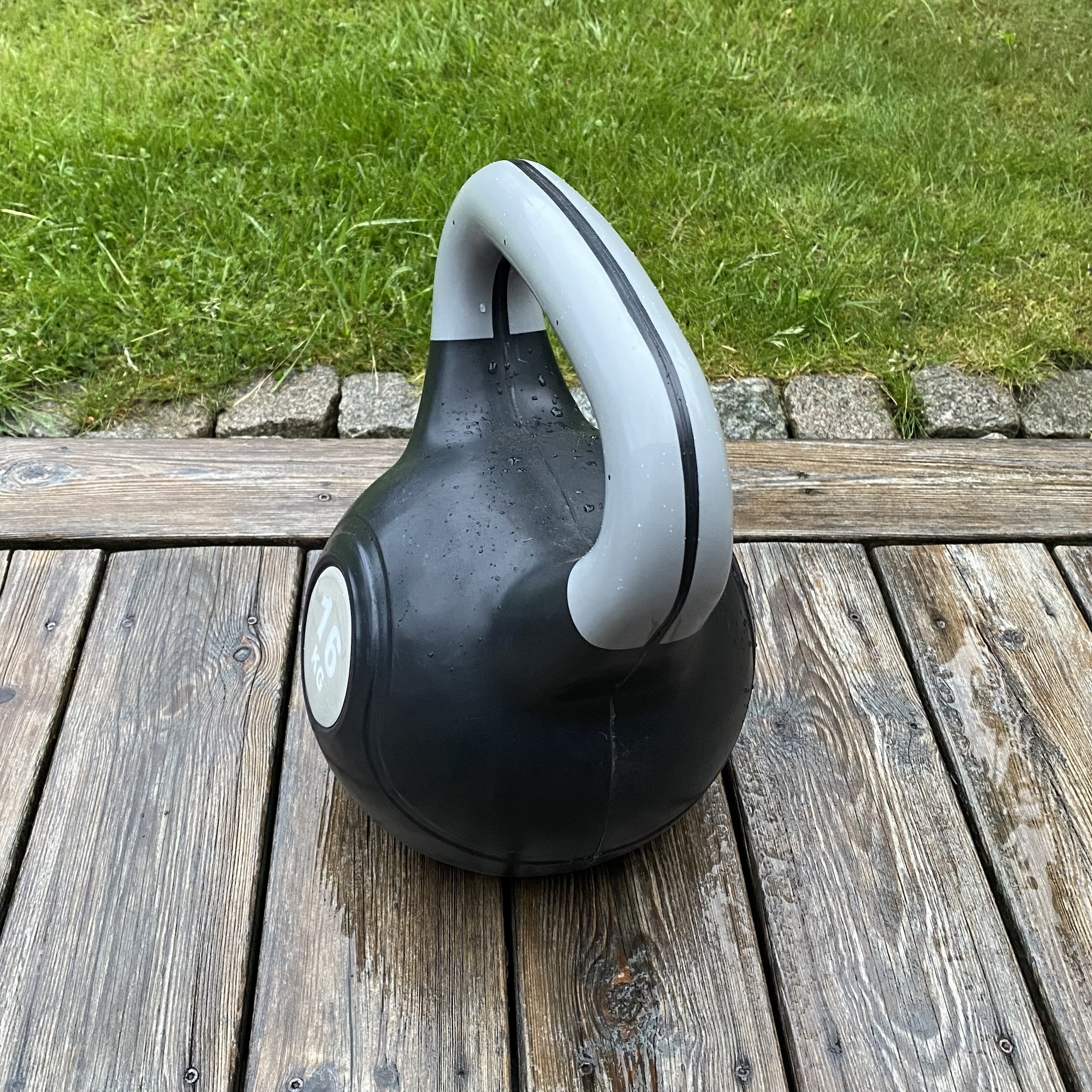 A black and grey kettlebell rests on a wet wooden deck beside grass after rain, matching the rhythm and English summer mood of the surrounding post.