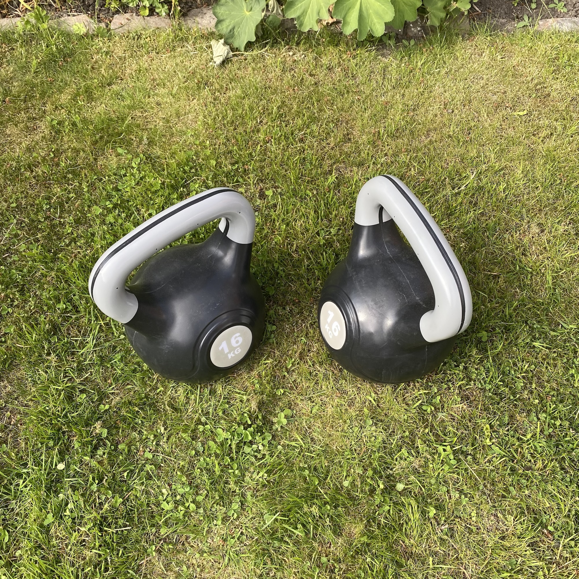 Two black 16 kg kettlebells rest on a grassy lawn, with white handles and weight labels visible beside garden edging and greenery.