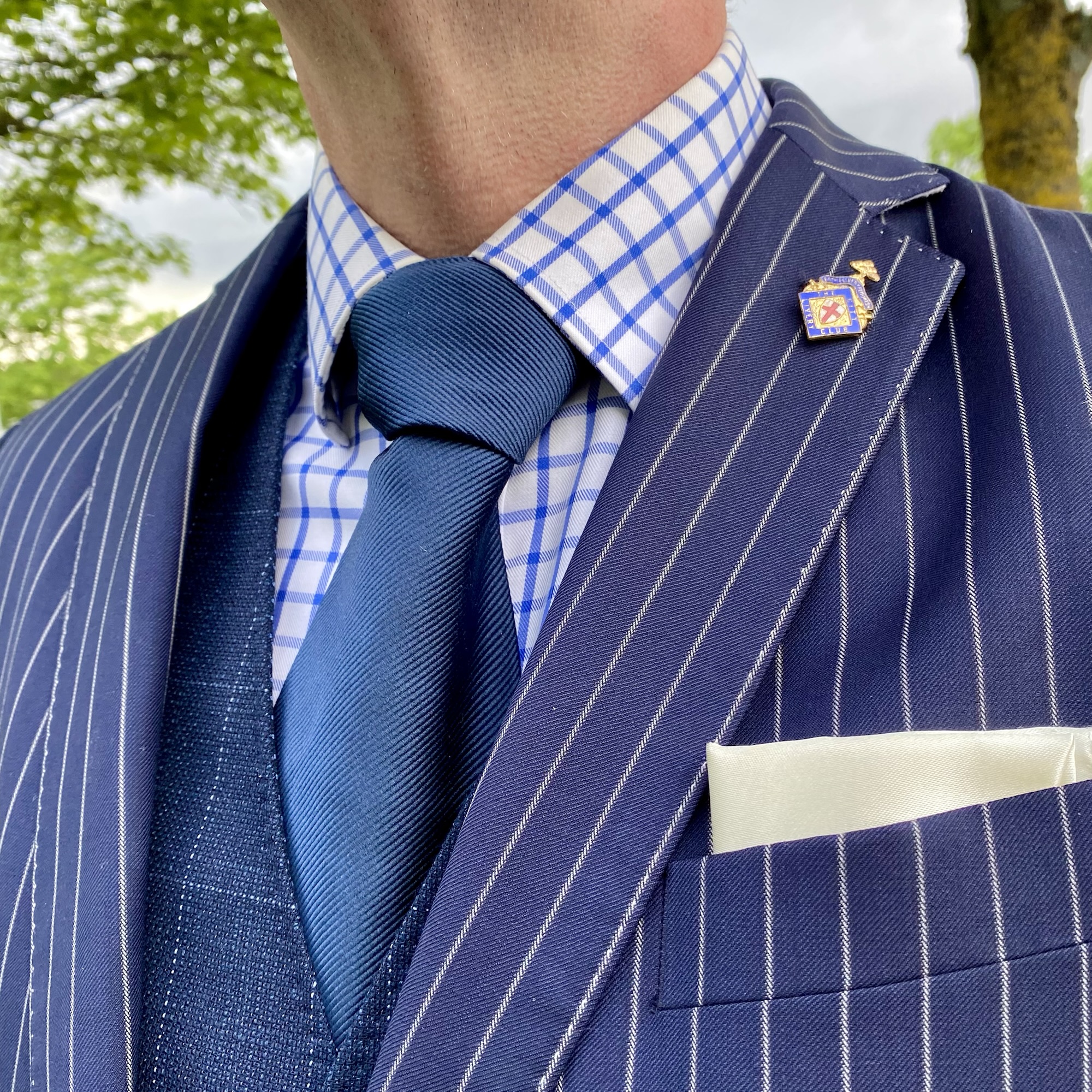 Blue tie and checked shirt under a navy pinstripe suit with a lapel pin, pocket square and blue-and-white checks beside blue stripes.