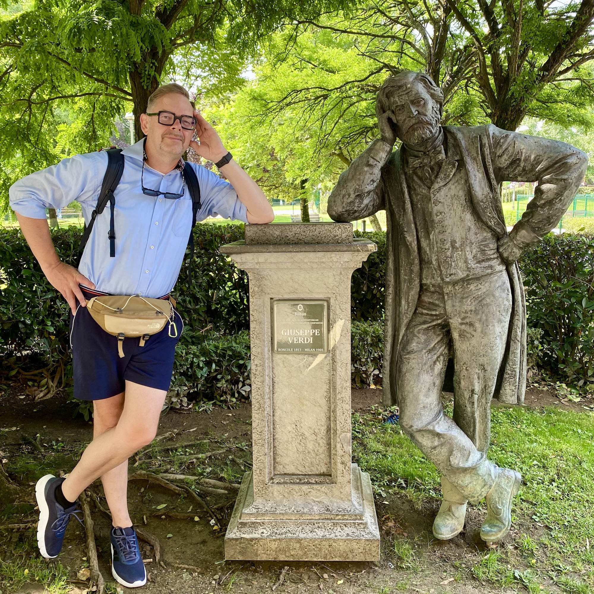 In a leafy Bilbao park, a man poses beside the Giuseppe Verdi statue, with the inscription visible on the pedestal.