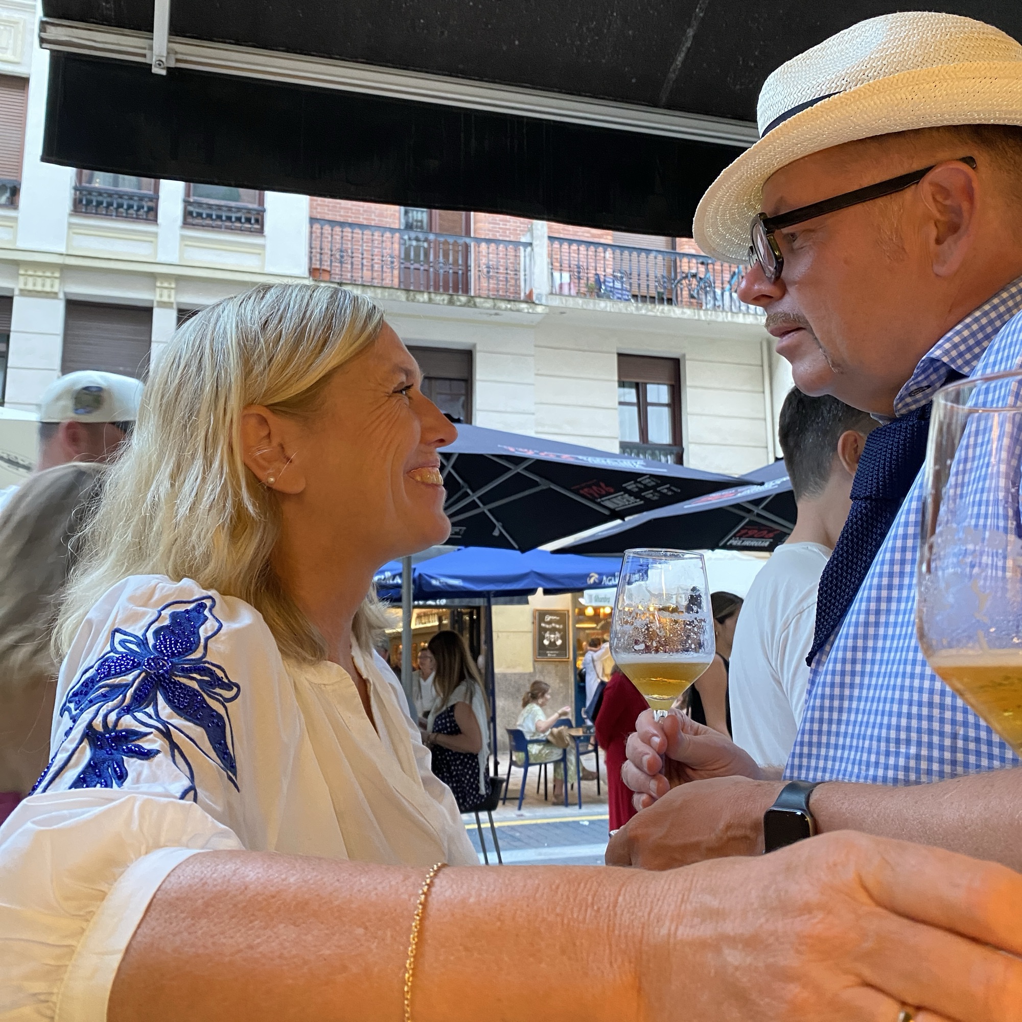 A blonde woman smiles closely at a man in a straw hat while holding a beer in a lively outdoor café.