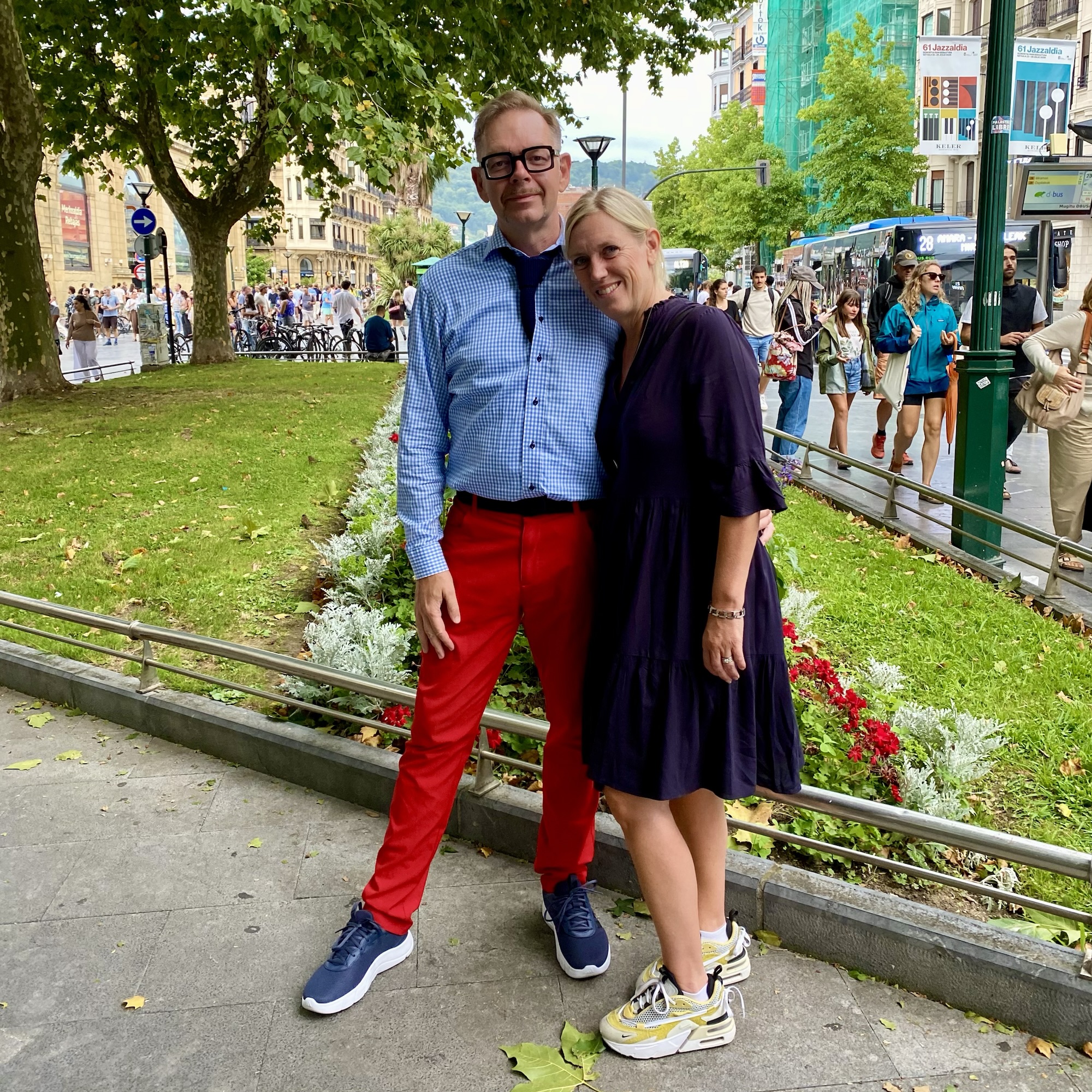 A man in red trousers and a woman in a navy dress stand on a busy, tree-lined European avenue with buses and passers-by.