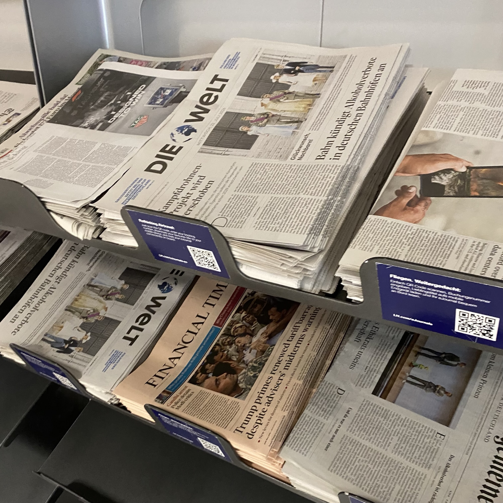 Stacks of printed German newspapers sit in a rack, with bold headlines and open pages showing articles and photographs.
