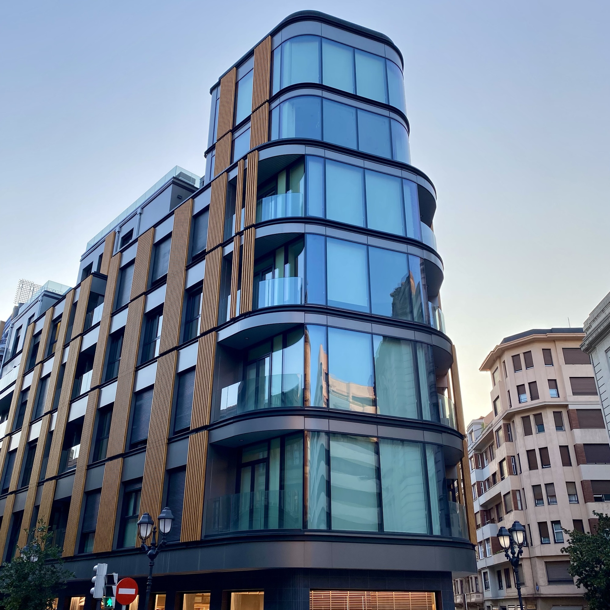 Modern curved glass building in Bilbao with dark cladding and vertical wooden slats beside older apartment blocks.
