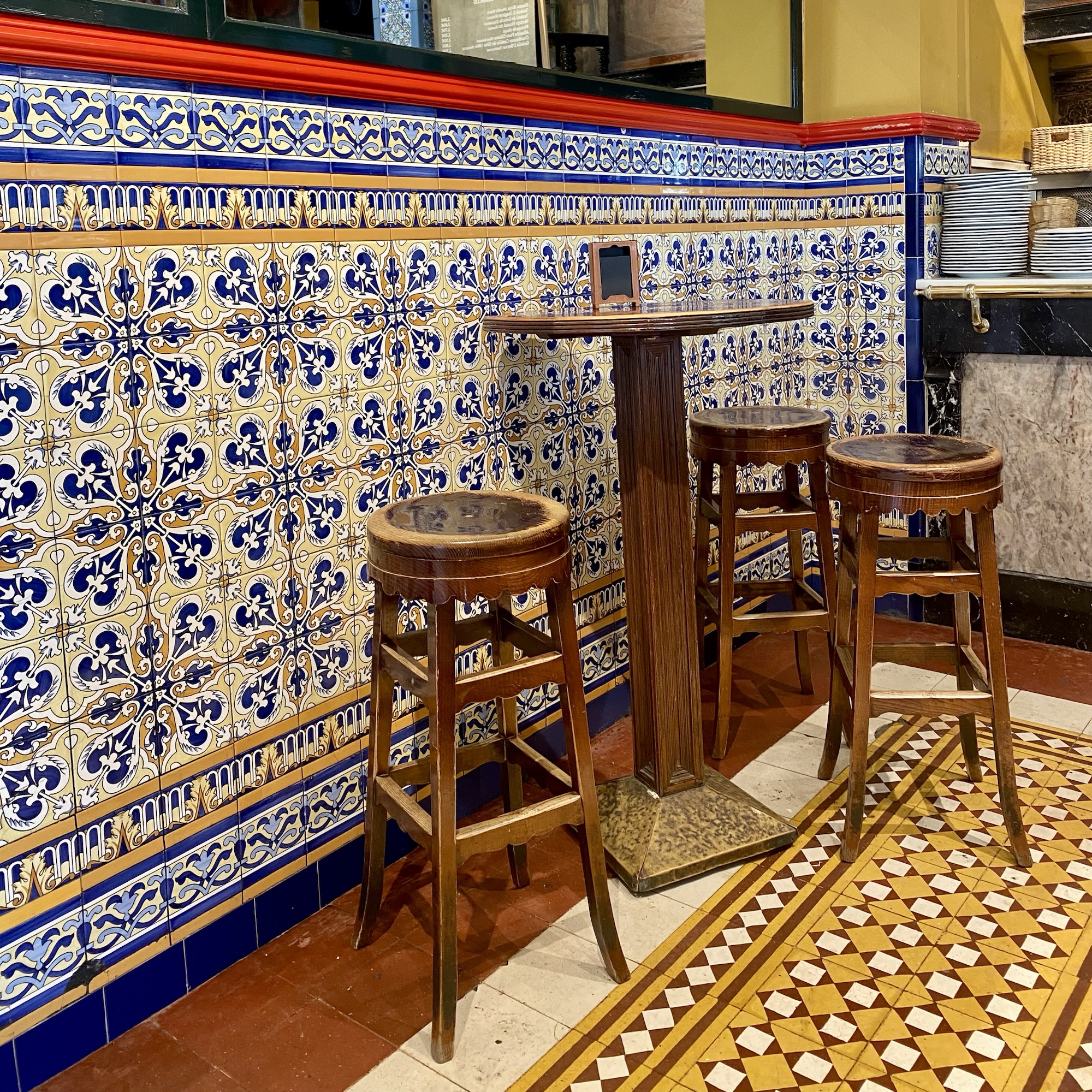 Ornate blue-and-yellow tiled walls frame a small café corner with wooden stools and a high table in Bilbao.