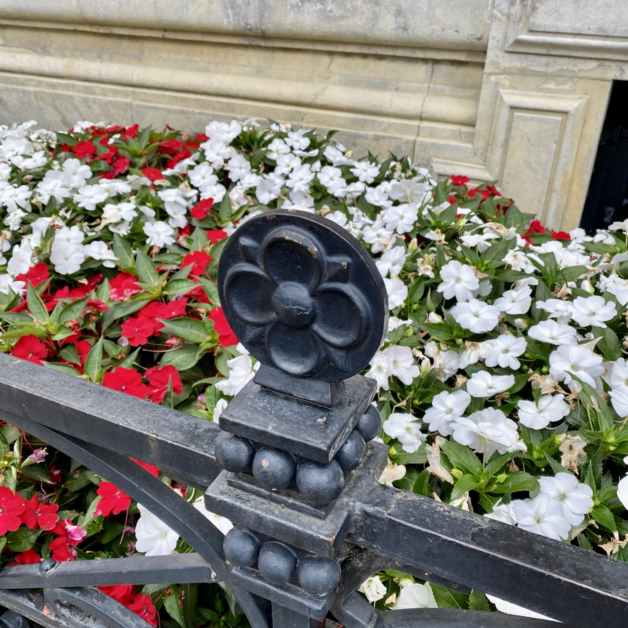 A black metal quatrefoil finial stands among red and white flowers beside a weathered stone wall.